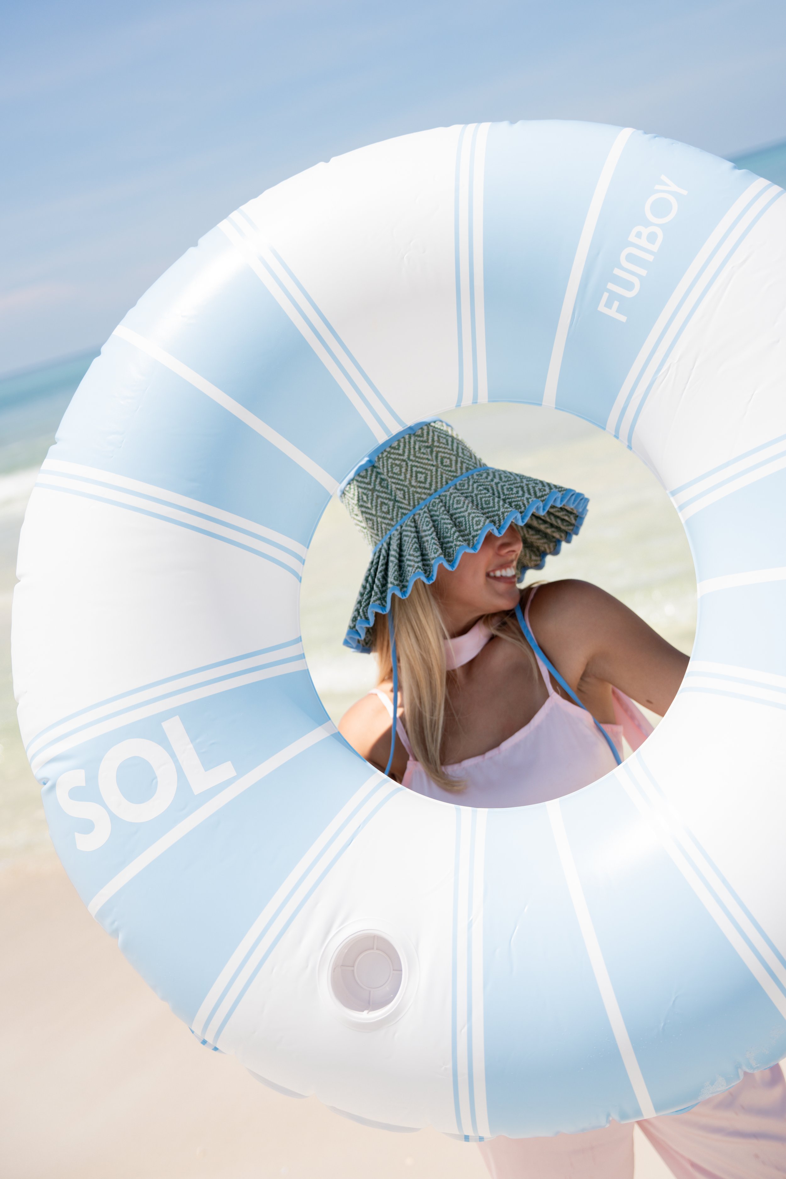 Woman on the beach holding a large inflatable pool float with blue and white stripes, and the words 'SOLAR' and 'FUBOY' printed on it. She is wearing a patterned towel or hat over her head, and a pink dress with thin straps. The background features t