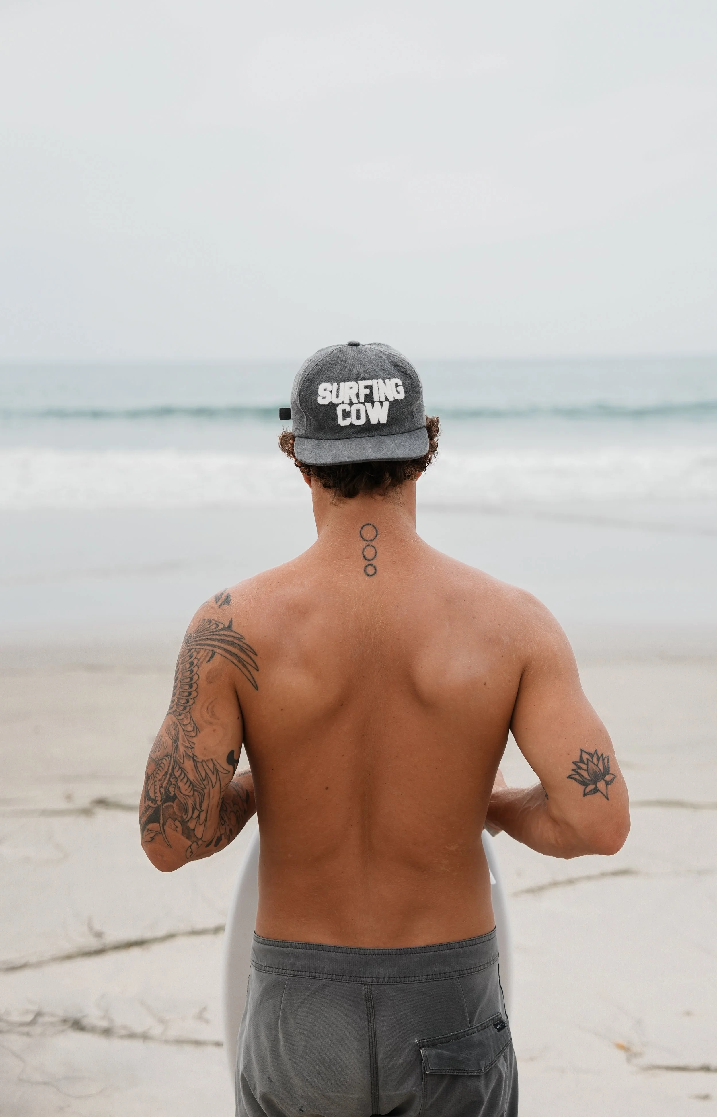 Back view of shirtless man with tattoos on arms and neck, wearing a gray cap that says 'Surfing Cow', standing at the beach facing the ocean on a cloudy day.