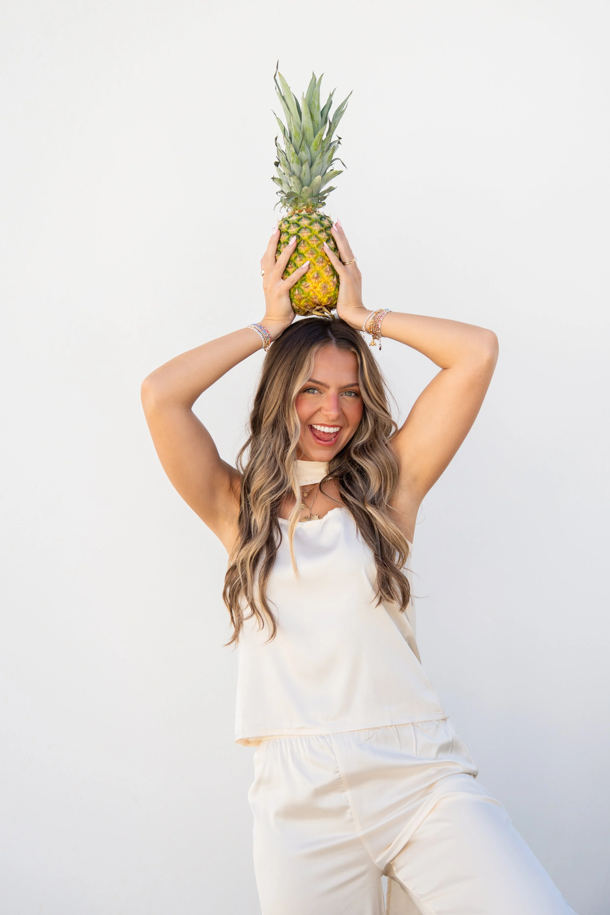 A woman with wavy light brown hair holding a pineapple above her head, smiling, against a plain white background.