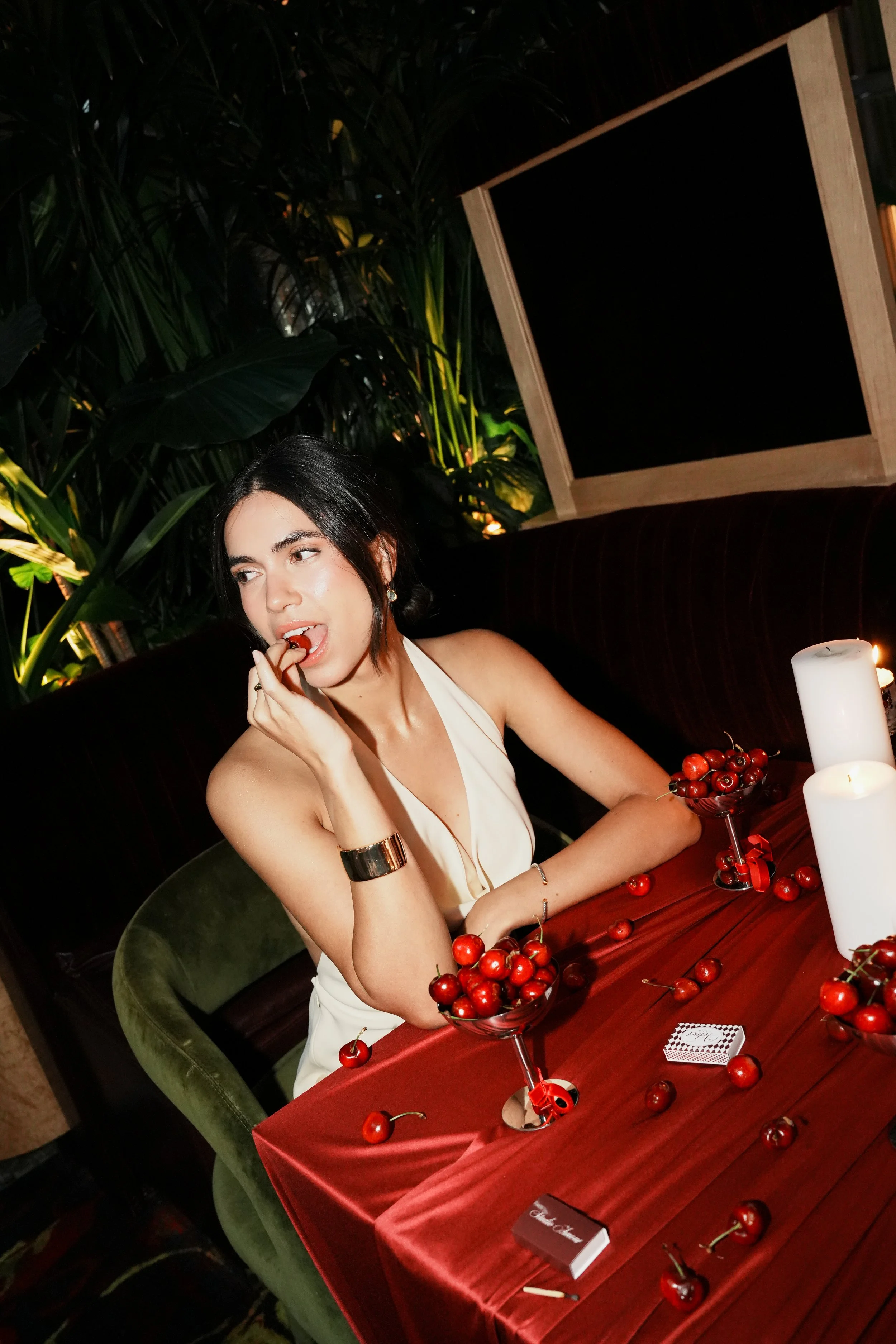 A woman with short dark hair and fair skin sitting at a table with a red tablecloth, surrounded by bowls of cherries and lit candles. She is wearing a white sleeveless dress and has jewelry on her wrist and ear. The background includes lush green pla