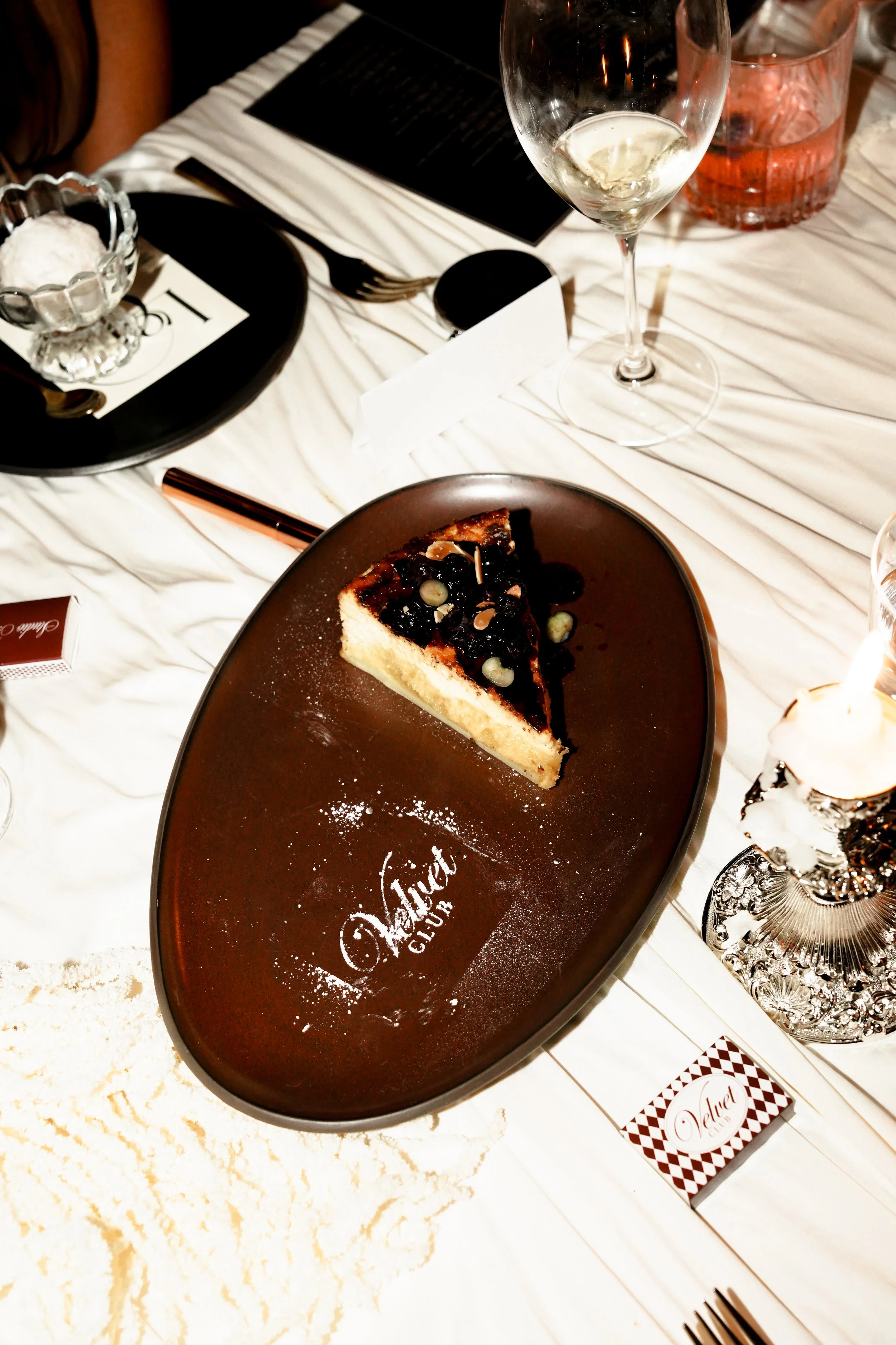 A dessert plate with a slice of cheesecake topped with dark berry sauce and garnished with sliced almonds, on a velvet-colored serving tray. A glass of white wine, a glass of pink-colored drink, and various silverware and tableware are on a white tab