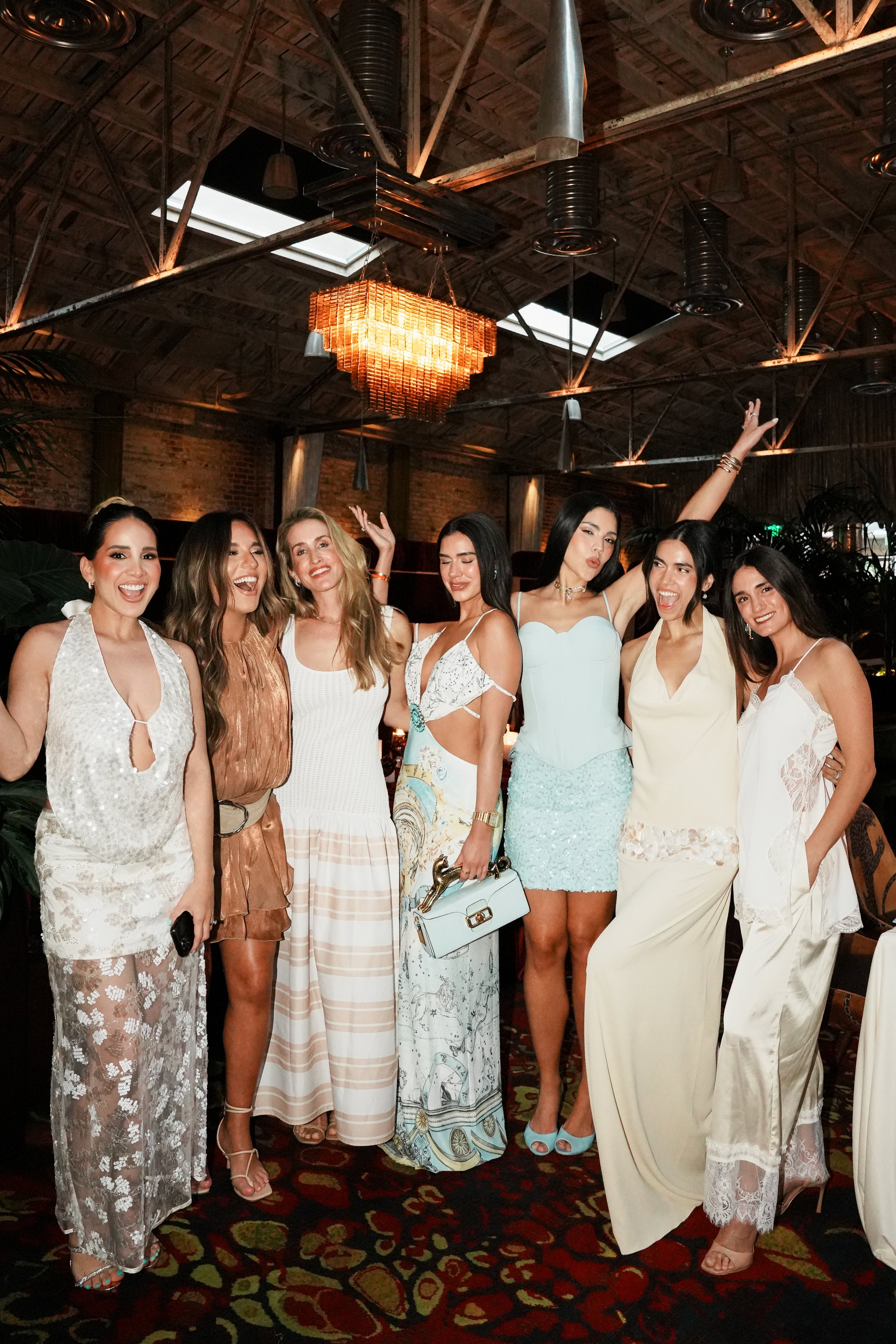A group of seven women dressed in stylish summer dresses standing together in a dimly lit indoor setting with exposed brick walls, wooden ceiling beams, and a decorative chandelier overhead. They are smiling and posing for the photo, showcasing their