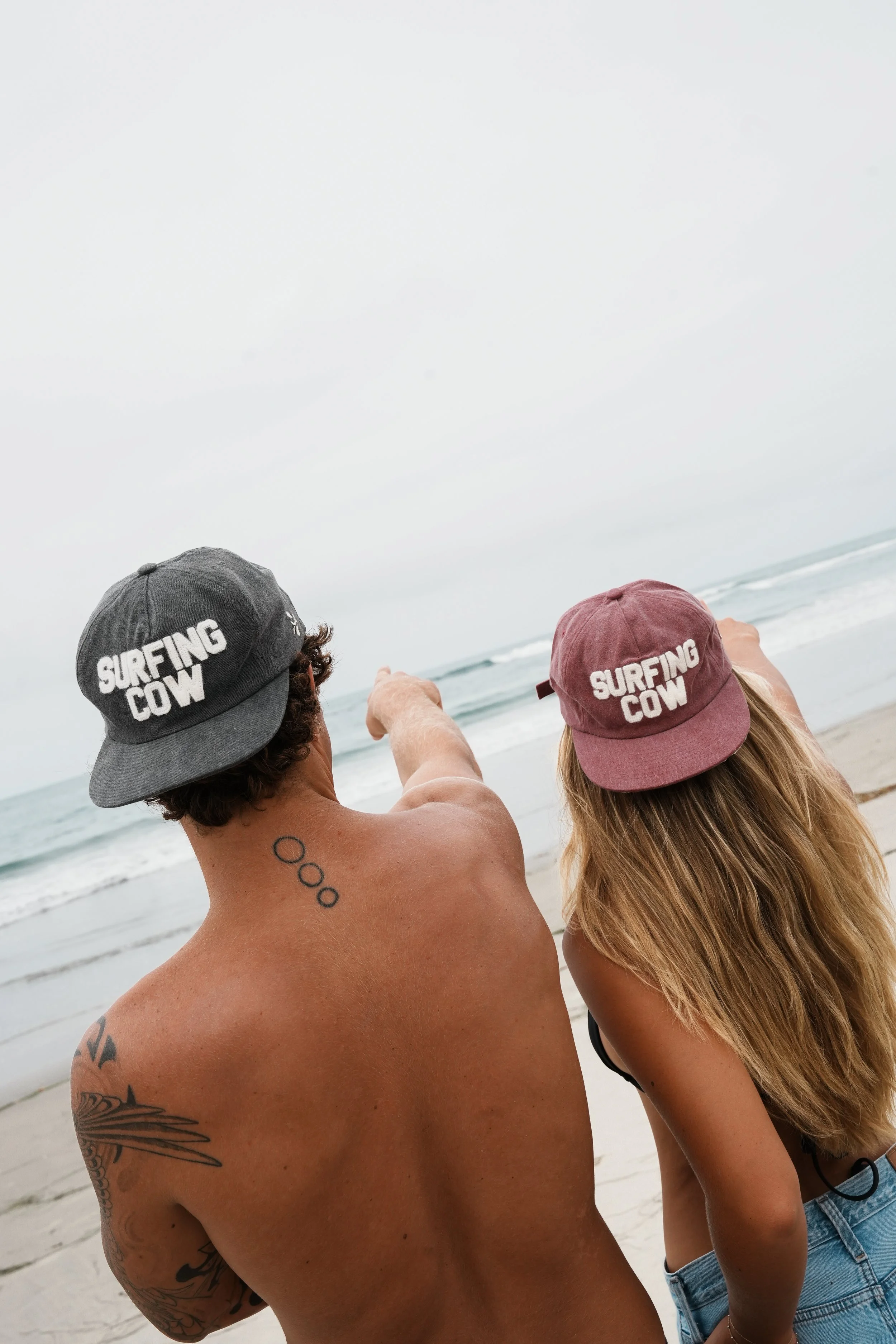 Two people, a shirtless man and a woman, are at the beach wearing hats that say "Surfing Cow." The man has tattoos on his arm and back, and the woman has long blonde hair and is wearing a black top and denim shorts. They are pointing towards the ocea