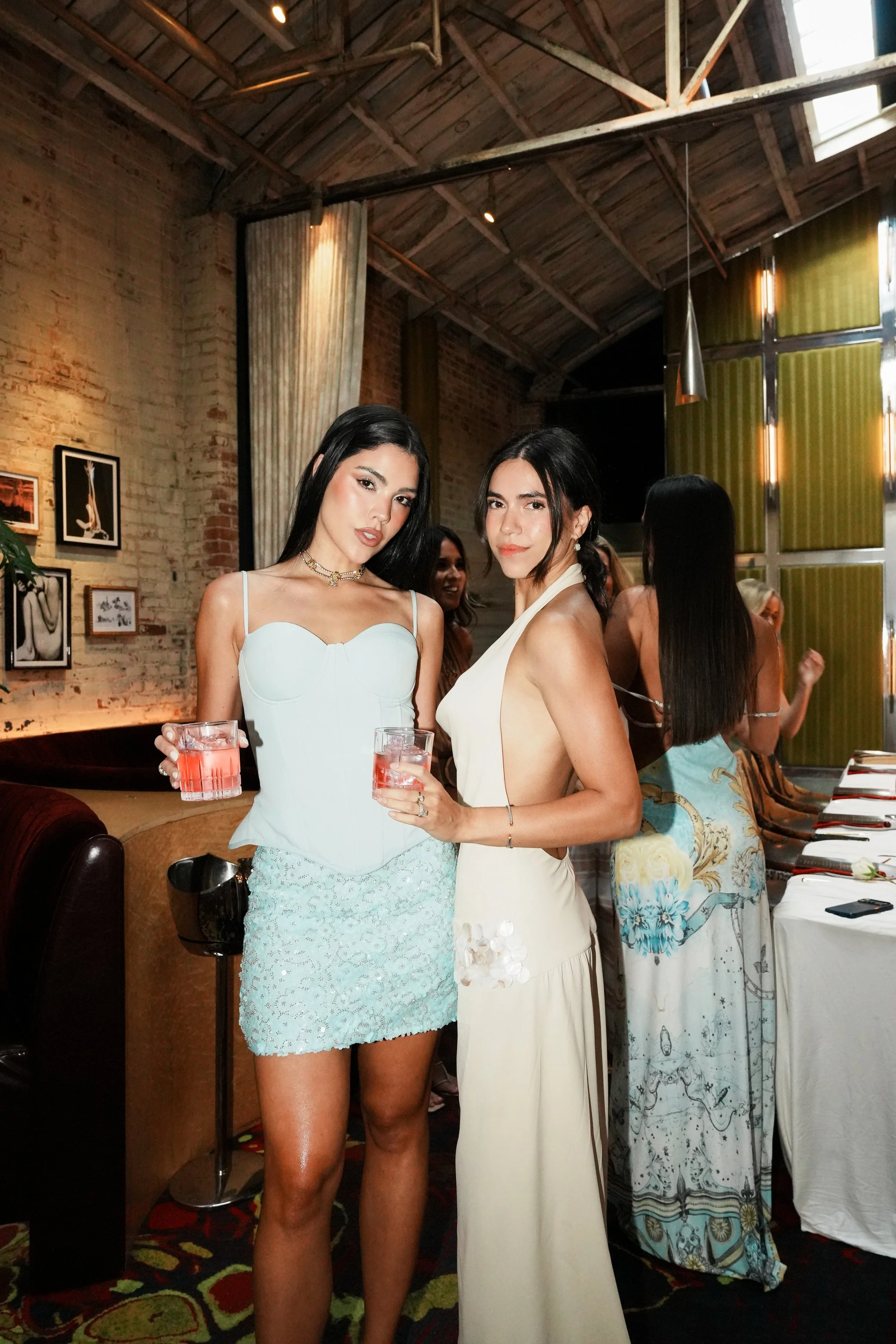 Two women dressed in elegant outfits holding drinks at a social gathering inside a rustic-style venue with exposed brick walls, framed artwork, and low lighting.
