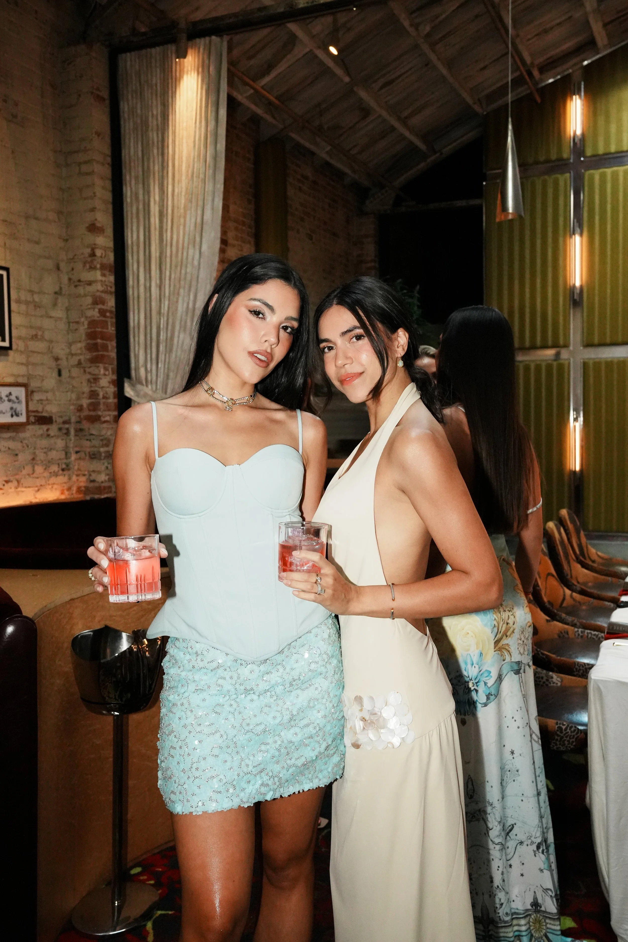 Two women in formal dresses standing side by side at a social gathering, holding drinks, smiling at the camera, in a warmly lit venue with brick walls and decorative curtains.