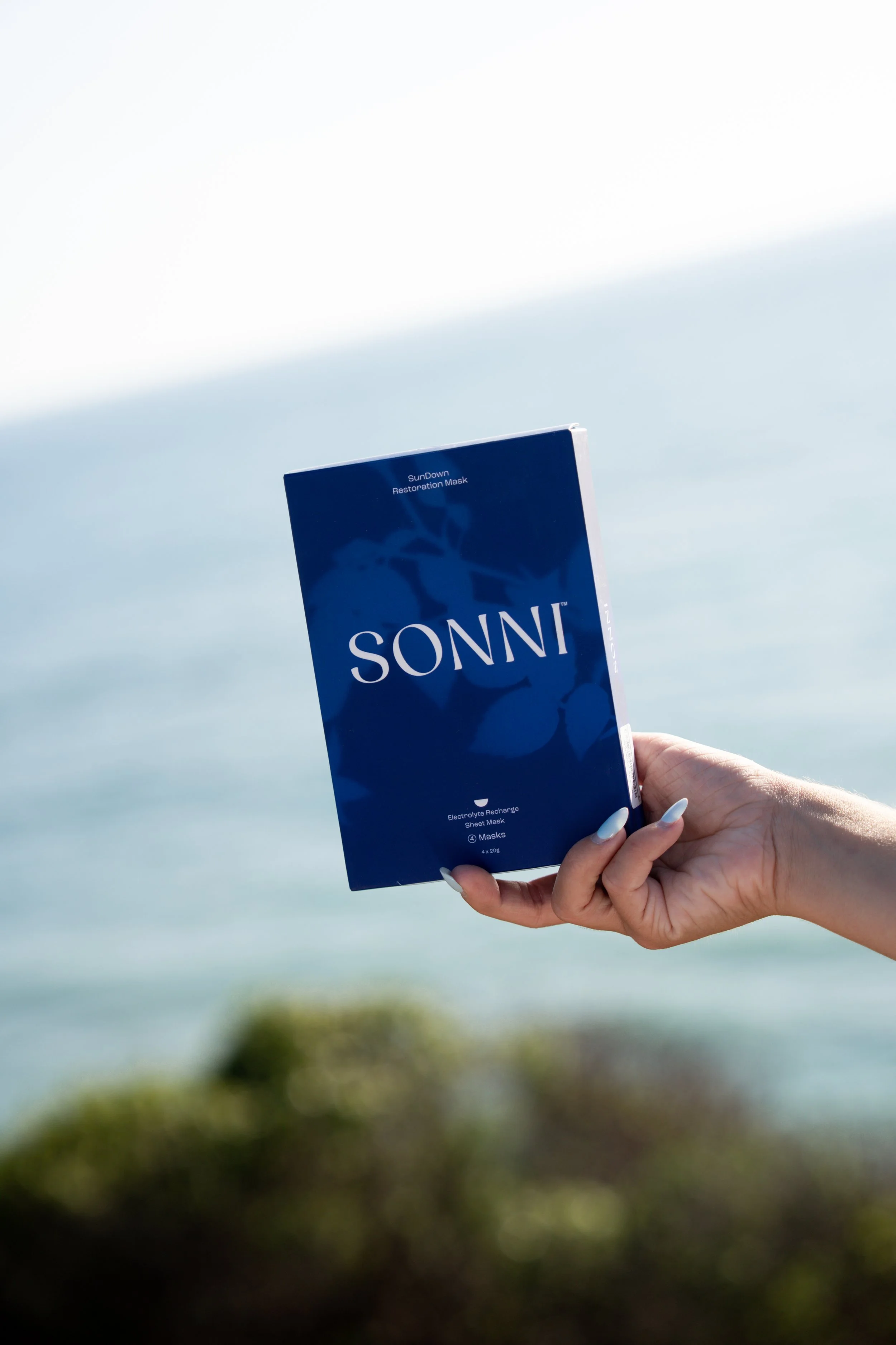 A person holding a blue package labeled 'SunDown Restoration Mask' against a blurred natural landscape background.