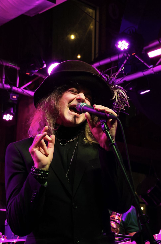 A woman singing into a microphone on stage, wearing a black hat and black blazer, with purple and pink stage lighting.