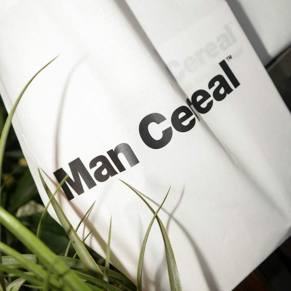 Close-up of a white bag with the logo and text 'Man Cereal' printed in black, partially obscured by green grass.