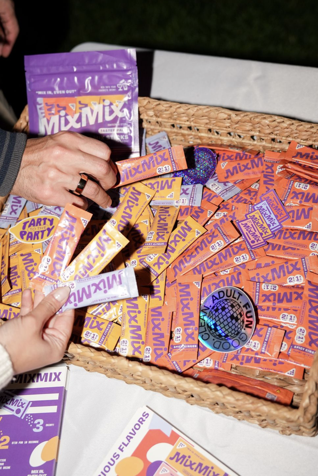 Two people, one adult and one child, reaching into a woven basket filled with orange and purple packages of MixMIX, a party candy, with a disc labeled 'Adult Fun' on top.
