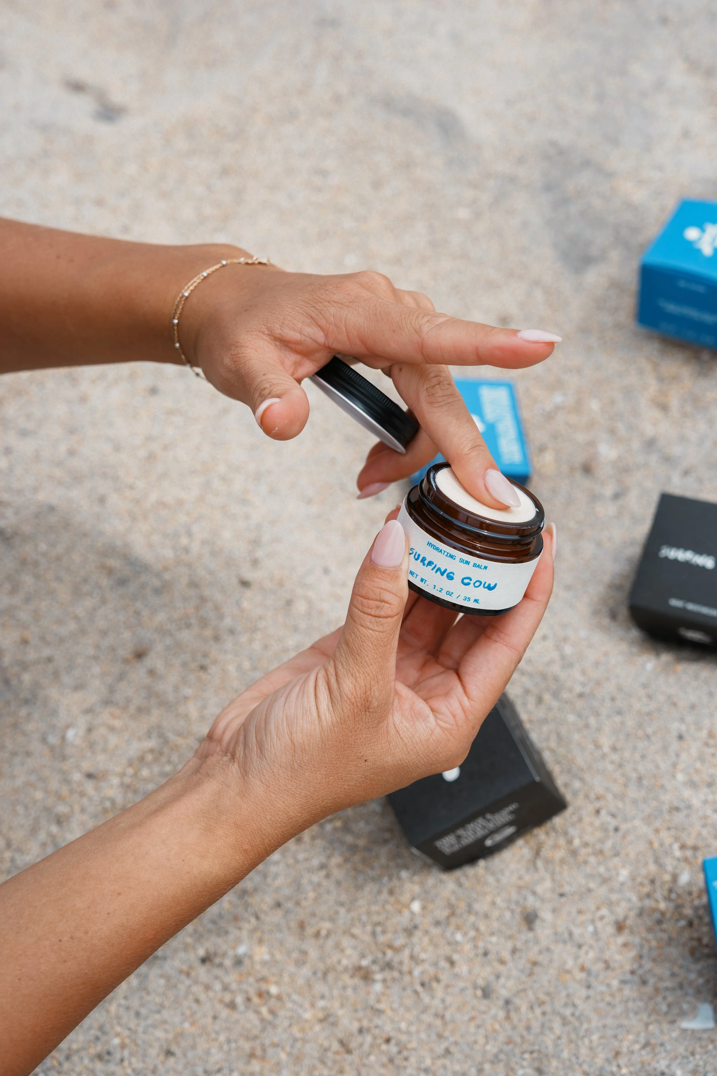 A person opening a jar of Surfing Cow hydrating sun balm on a textured surface outdoors.