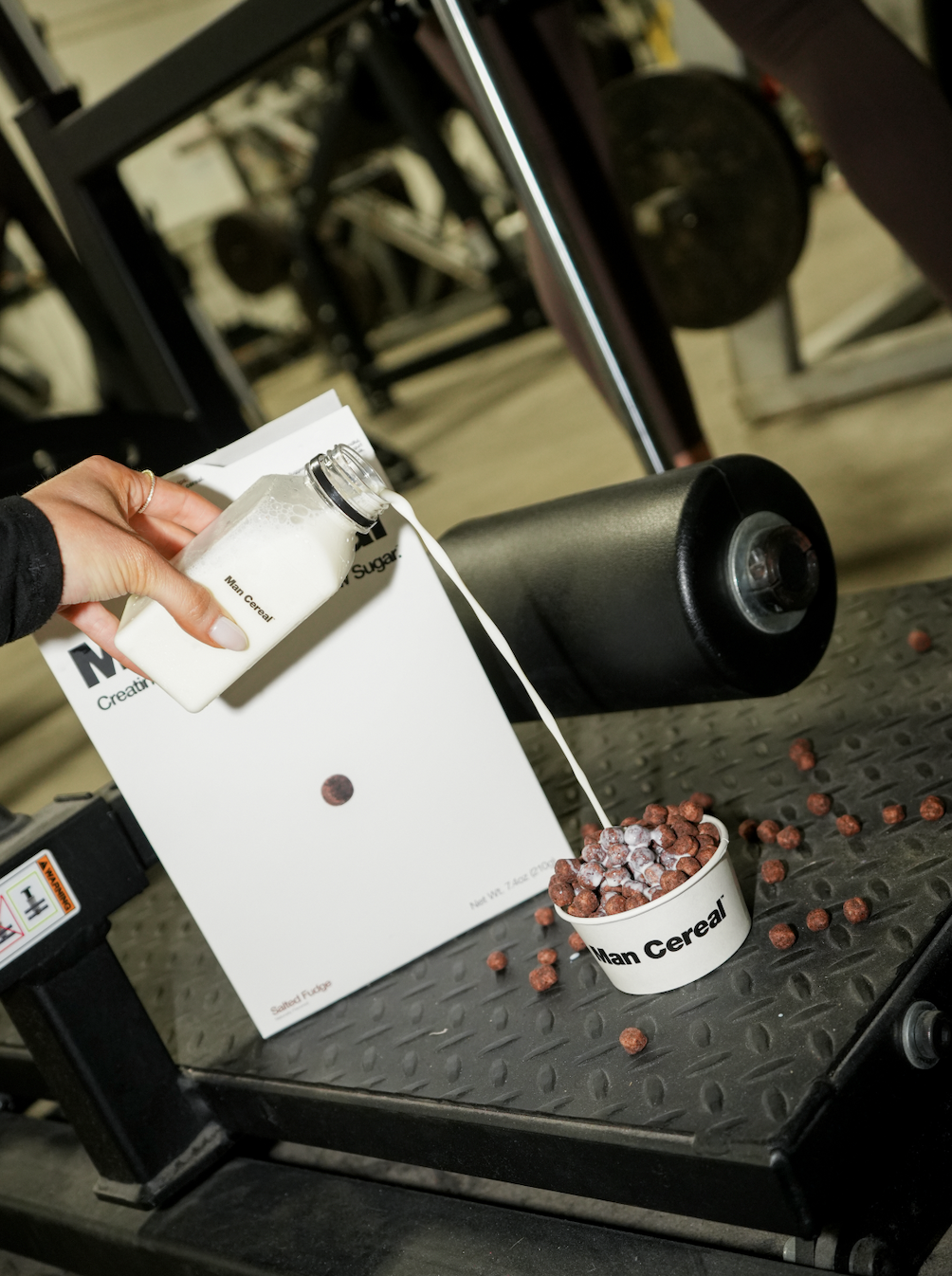Person pouring milk into a cereal bowl labeled 'Man Cereal' on a gym-industrial style black workout bench with scattered cereal pieces around.