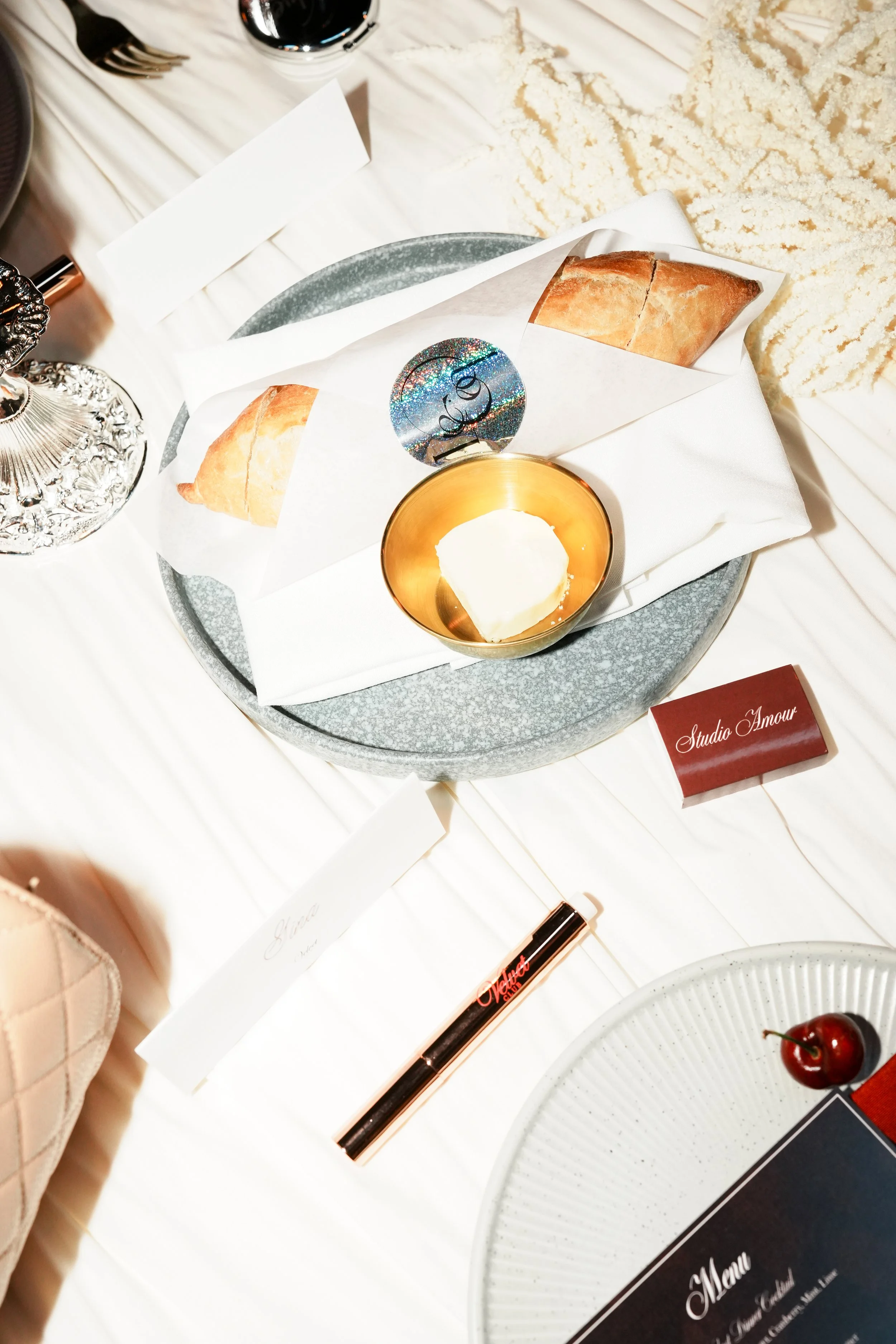 A tray with two paper packages of bread, a small container of butter, a cherry, a black lipstick tube, a white plate, and some decorative items on a white tablecloth.
