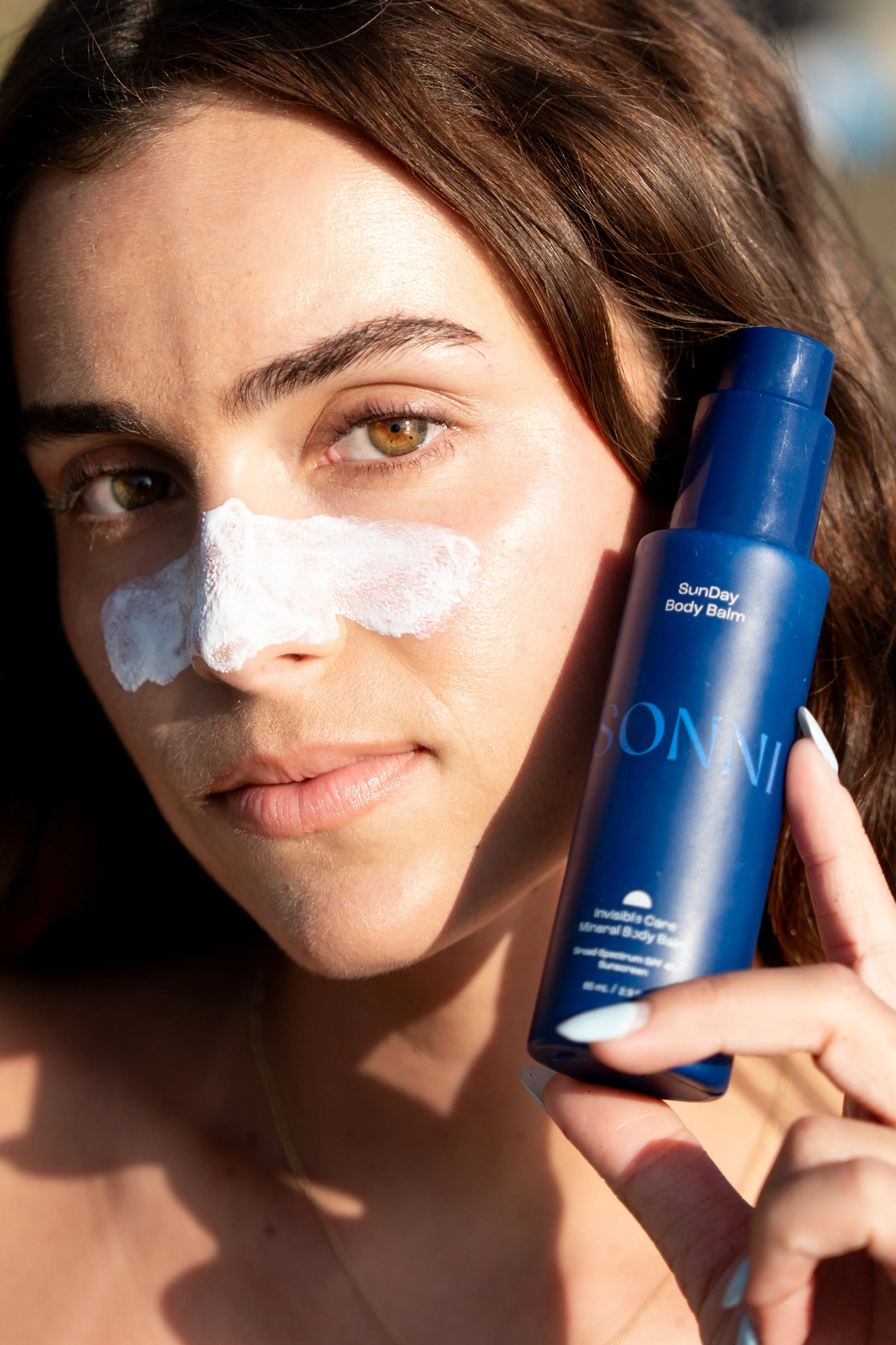 A woman with brown hair and hazel eyes applying white sunscreen to her nose while holding a blue bottle labeled 'SunDay Body Balm' in sunny outdoor setting.