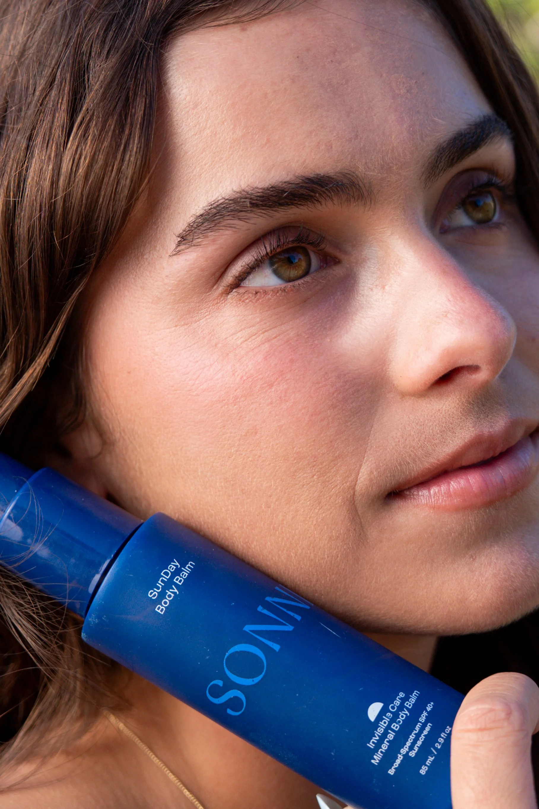 Close-up of a woman with brown hair, brown eyes, and natural makeup, holding a blue tube of sunscreens or skincare product against her cheek and jawline.