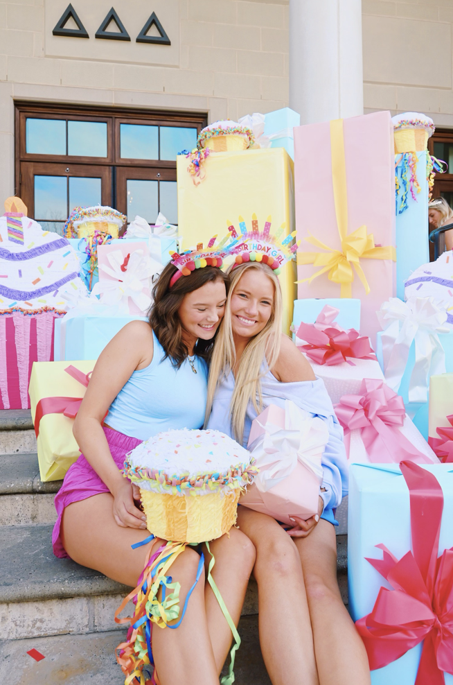 Two smiling young women sitting on steps surrounded by colorful birthday gifts, including large wrapped presents and a giant cupcake, celebrating a birthday party.