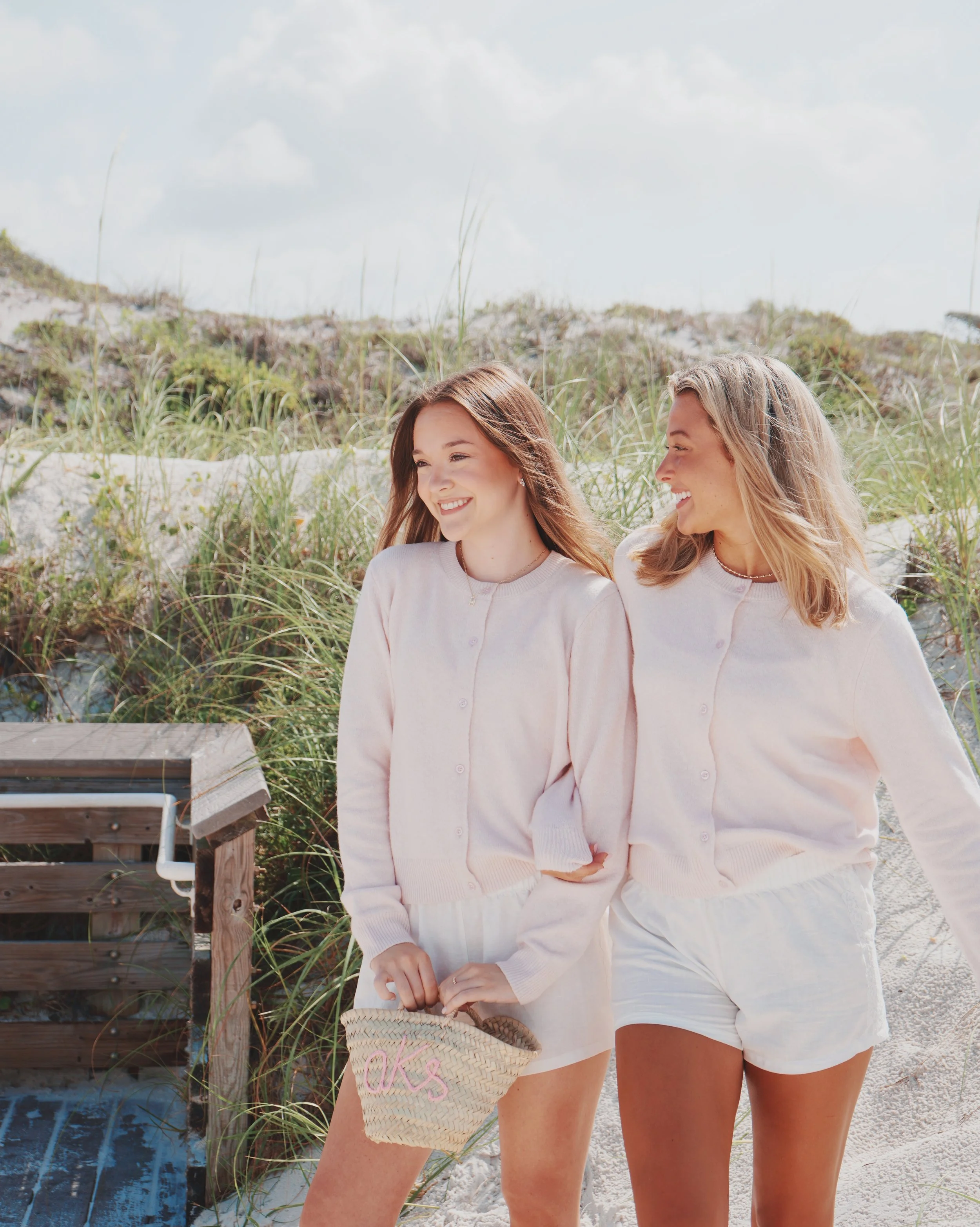 Two women smiling and walking on a sandy beach with seagrass and a wooden staircase in the background. They are wearing matching light-colored sweaters and shorts, and holding a straw bag with pink embroidery.