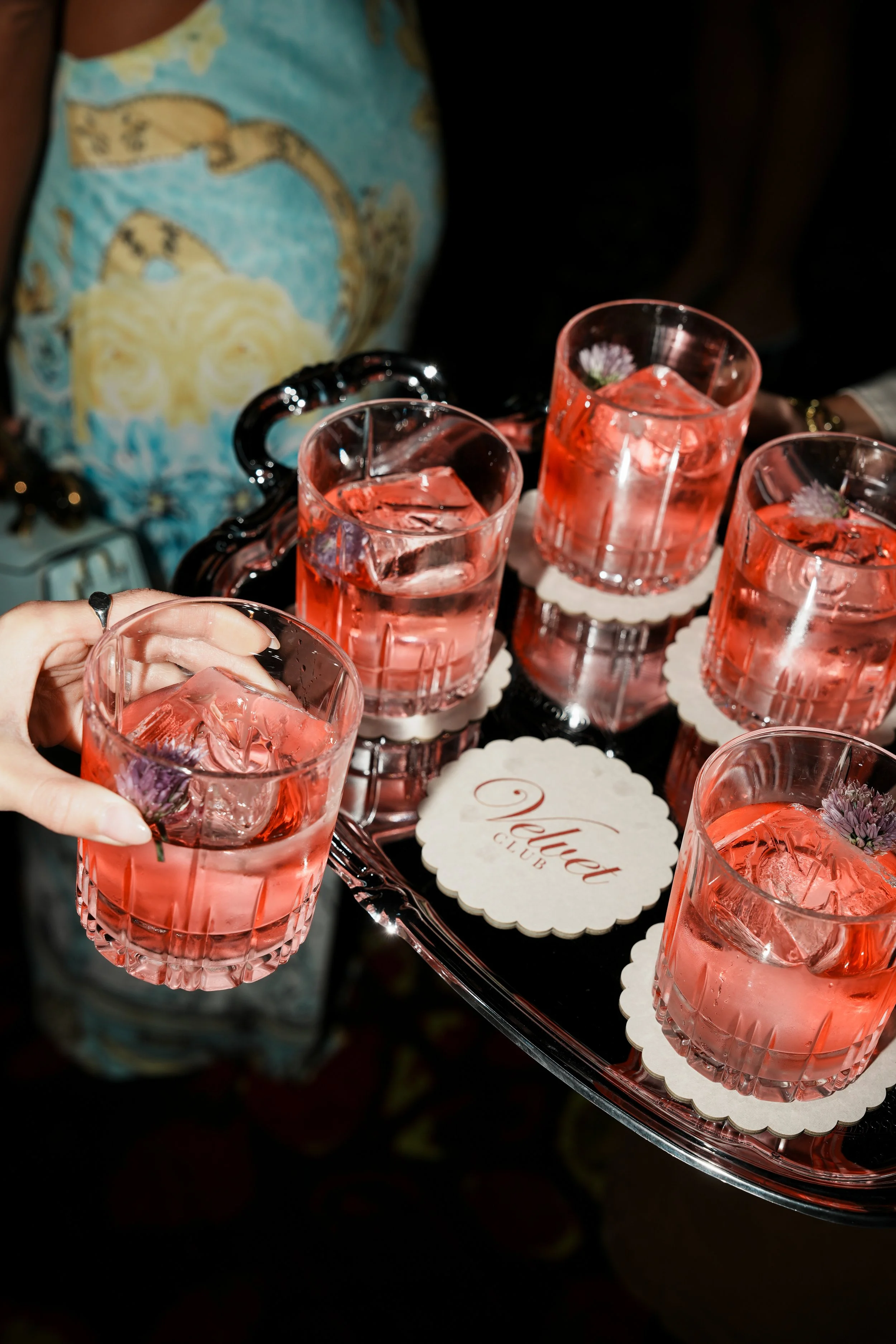 A person is holding a glass with pink drink and ice cubes, with several similar glasses on a tray, and a sign that reads 'Never Club' on a table.