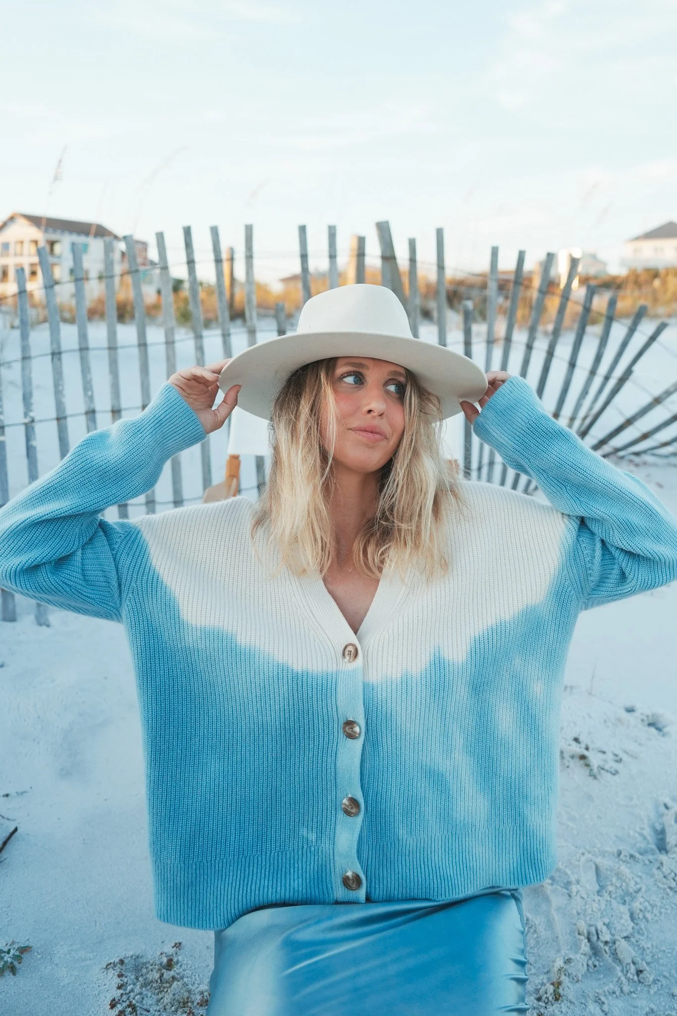 Young woman at the beach wearing a wide-brimmed white hat and a light blue cardigan, with a wooden fence and houses in the background.