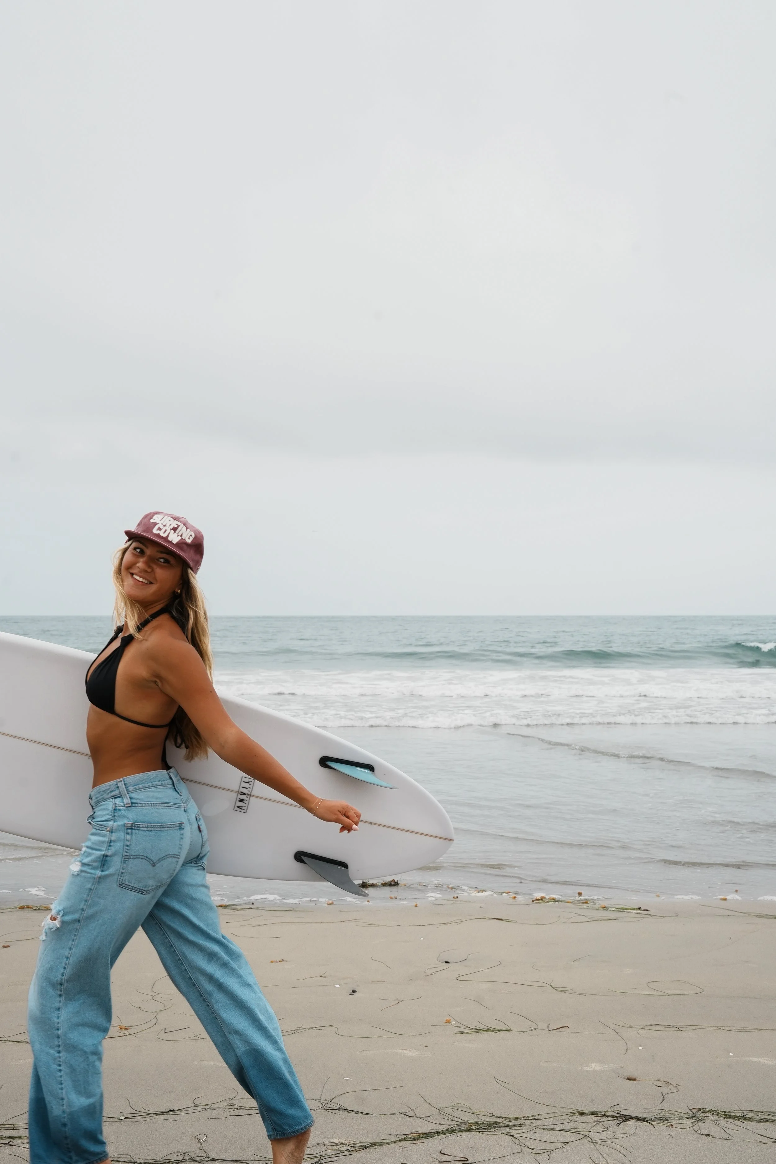 A woman standing on the beach holding a white surfboard under her arm, smiling; she is wearing a pink cap, black bikini top, and blue jeans with a rip on the right knee. The ocean waves and cloudy sky are visible in the background.