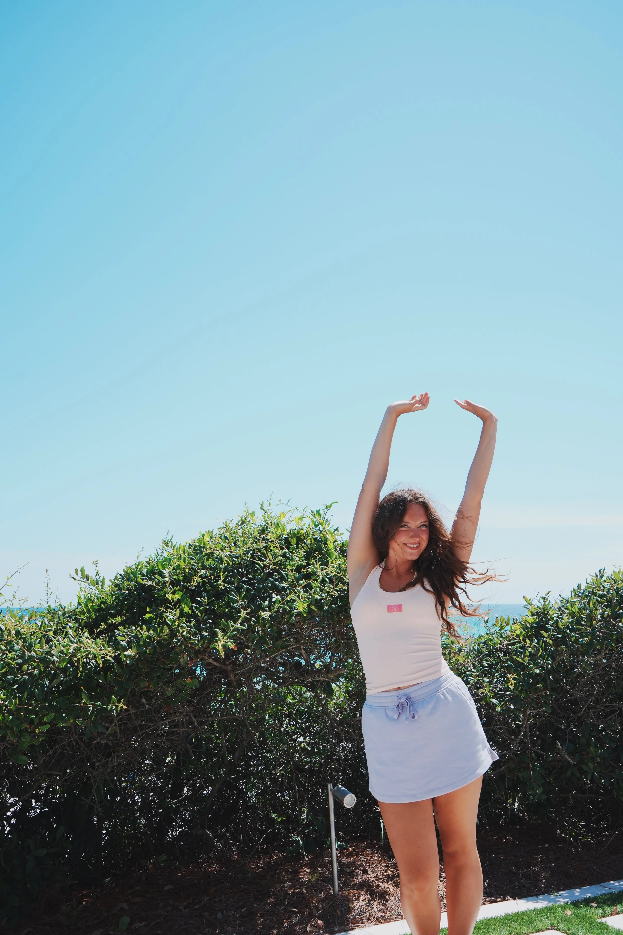 A smiling woman in a white tank top and white shorts standing outdoors with arms raised, bush and blue sky in background.
