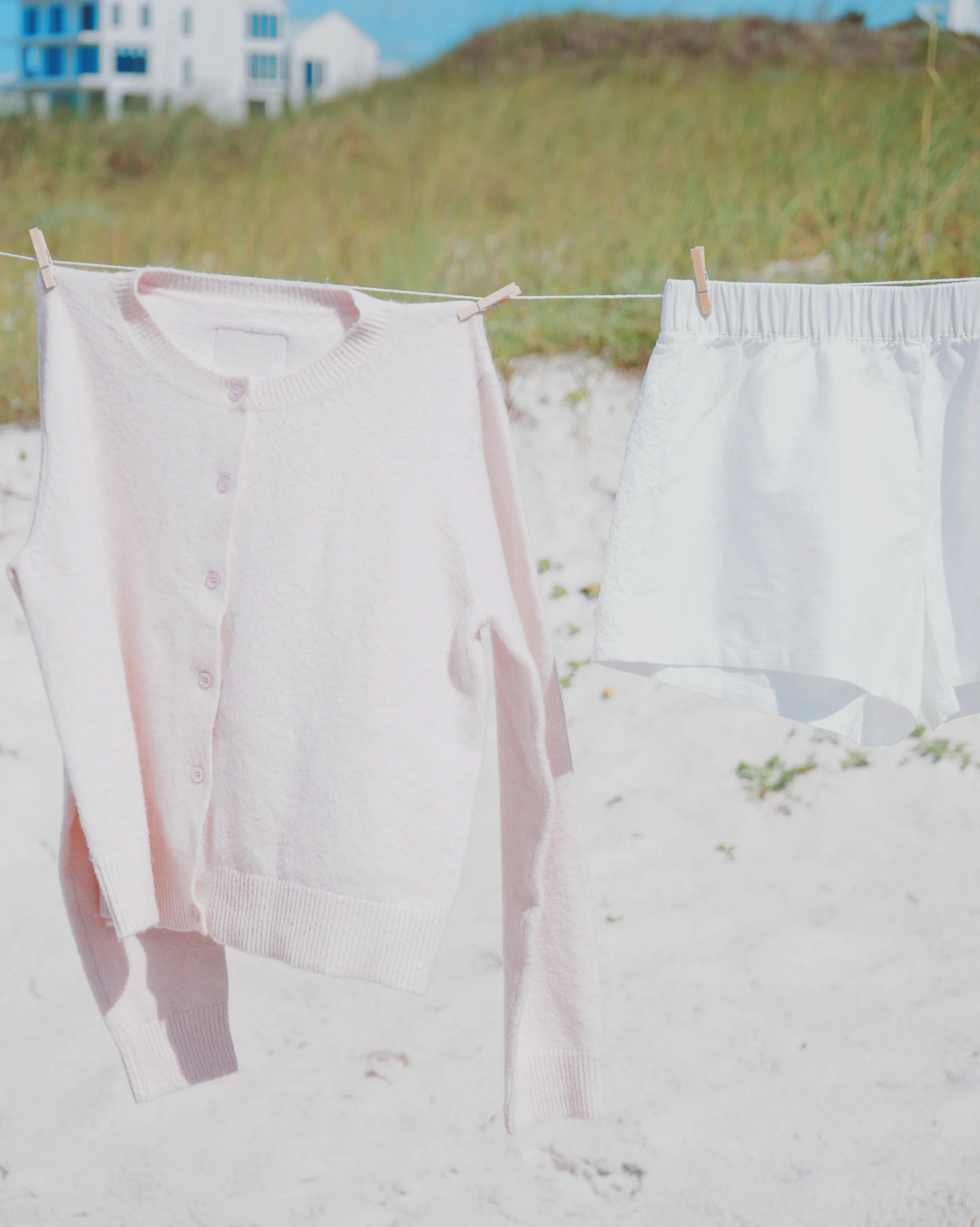 A pink cardigan and white shorts hanging on a clothesline with clothespins on a beach, with grassy dunes and a building in the background.