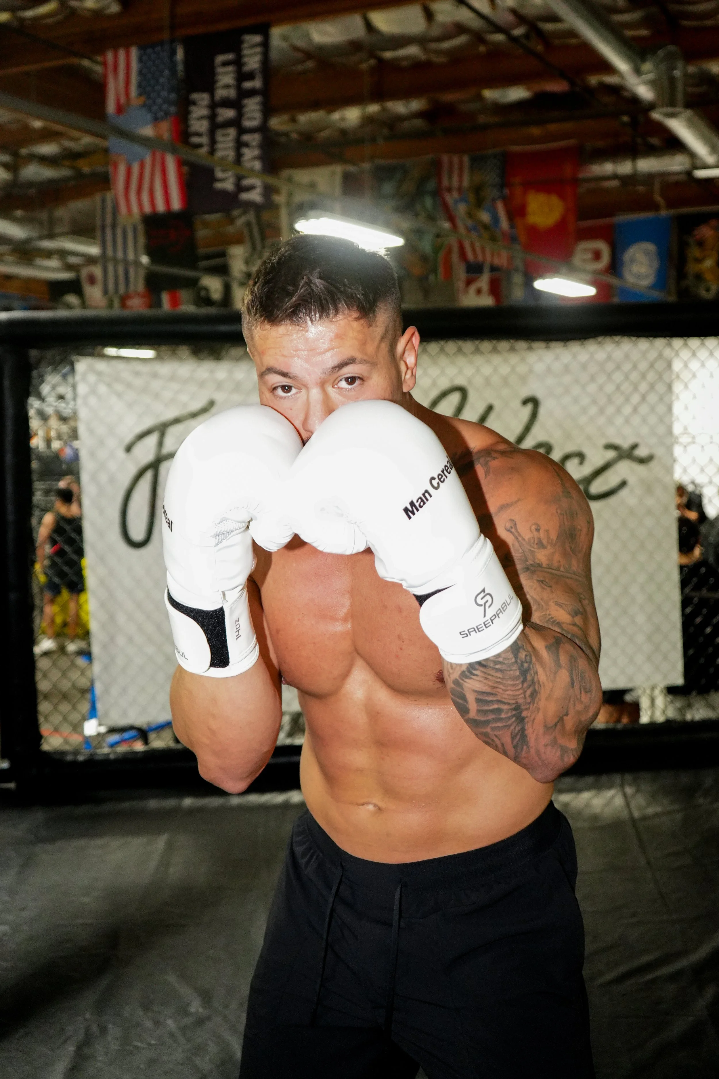 A shirtless male mixed martial artist wearing white MMA gloves poses in a fighting stance inside a training gym with various flags hanging from the ceiling.