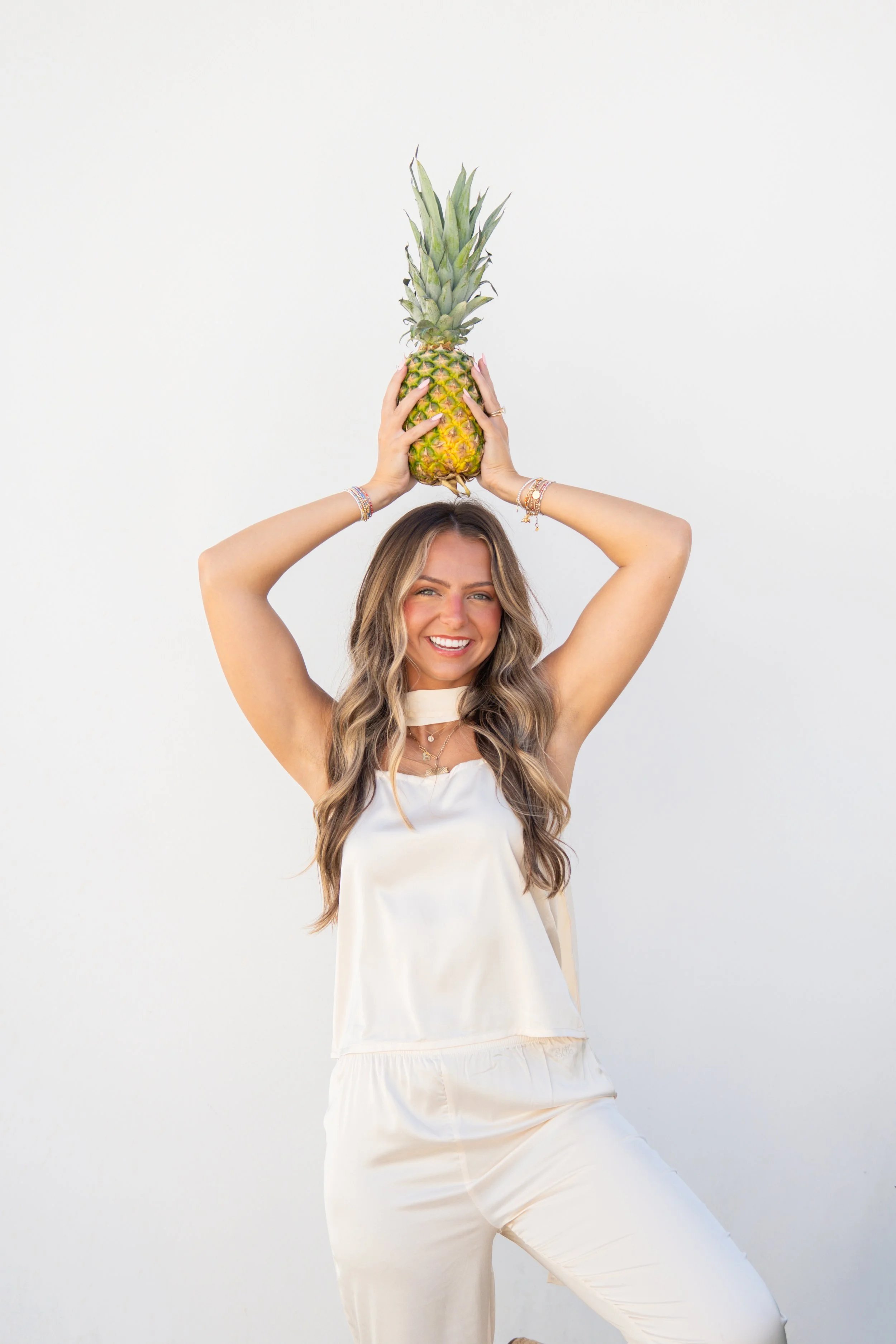 A woman smiling and dressed in white holds a pineapple above her head with both hands, standing against a plain white background.