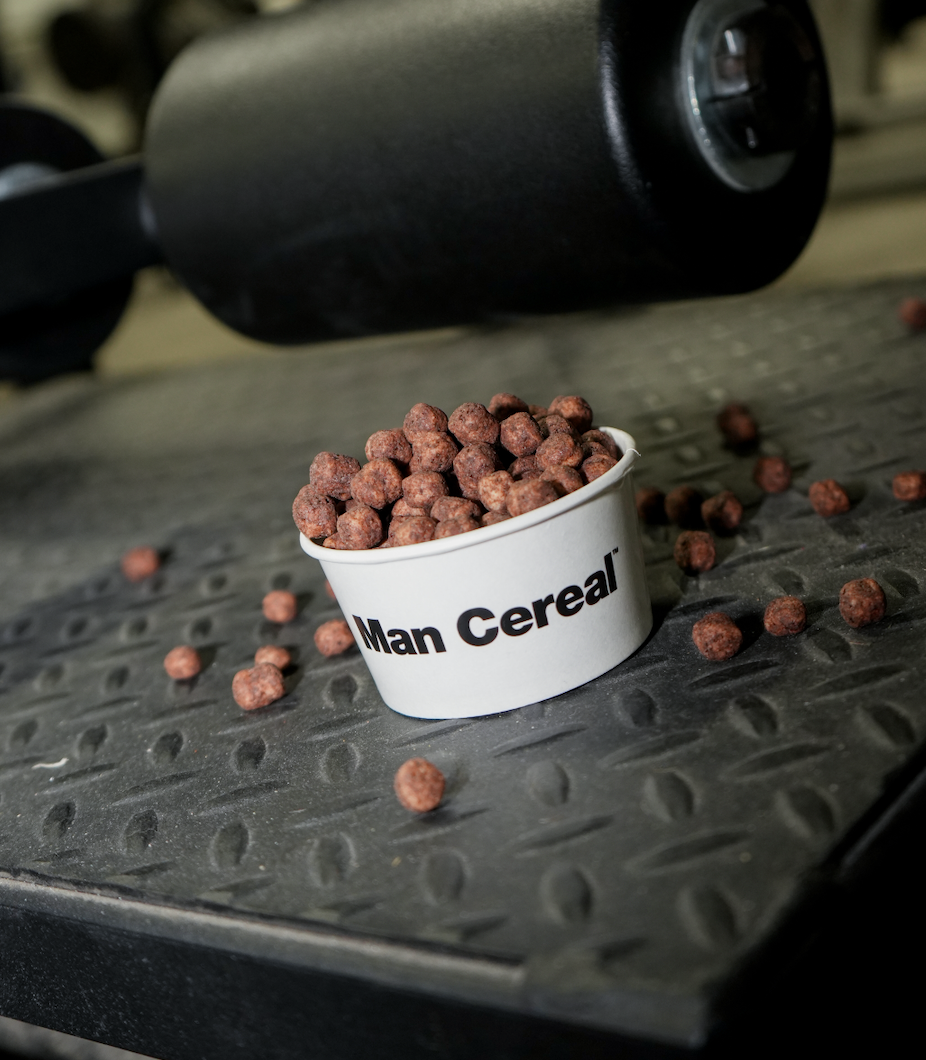 A container labeled 'Man Cereal' filled with chocolate cereal balls, spilled on a textured black surface, with some cereal pieces scattered around and part of a black object in the background.