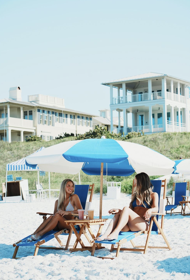Two women sitting on beach chairs under a large beach umbrella, talking and smiling, with drinks on a small table beside them, at a sunny beach.