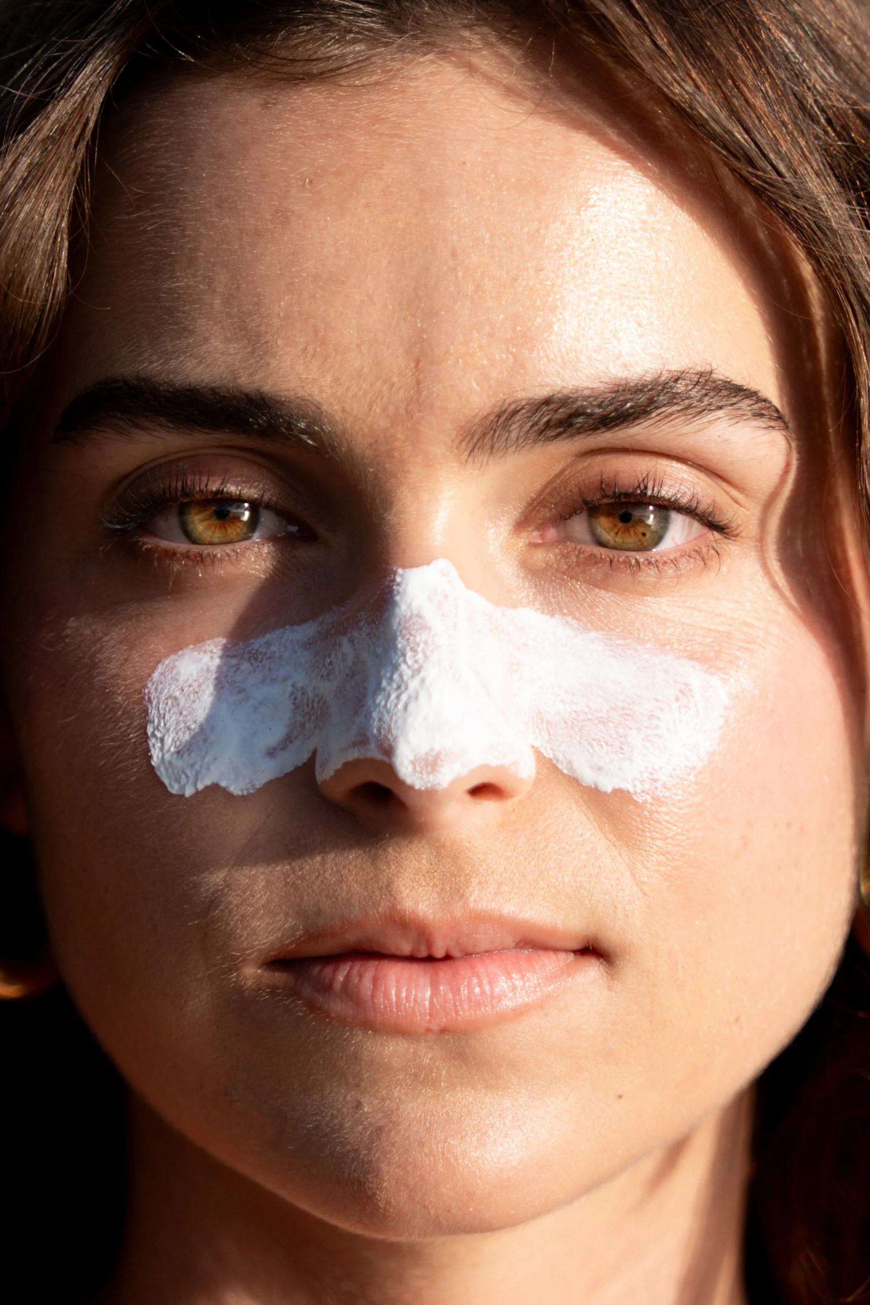 Close-up of a woman with brown hair and hazel eyes, wearing a white facial mask on her nose.