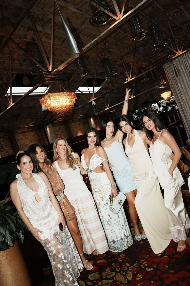 Group of seven women dressed in stylish summer and evening outfits posing together in a dimly lit, rustic indoor setting with exposed brick walls and wooden ceiling beams, decorated with chandeliers and industrial ductwork.