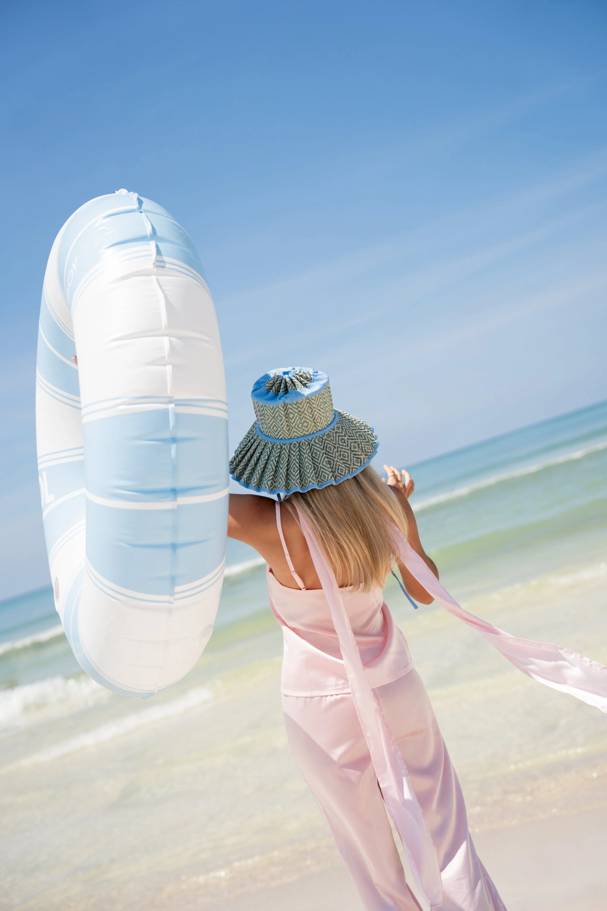 A woman with long blonde hair wearing a wide-brimmed hat, a pink cover-up, holding a large inflatable pool float, standing on a sandy beach with the ocean and blue sky in the background.