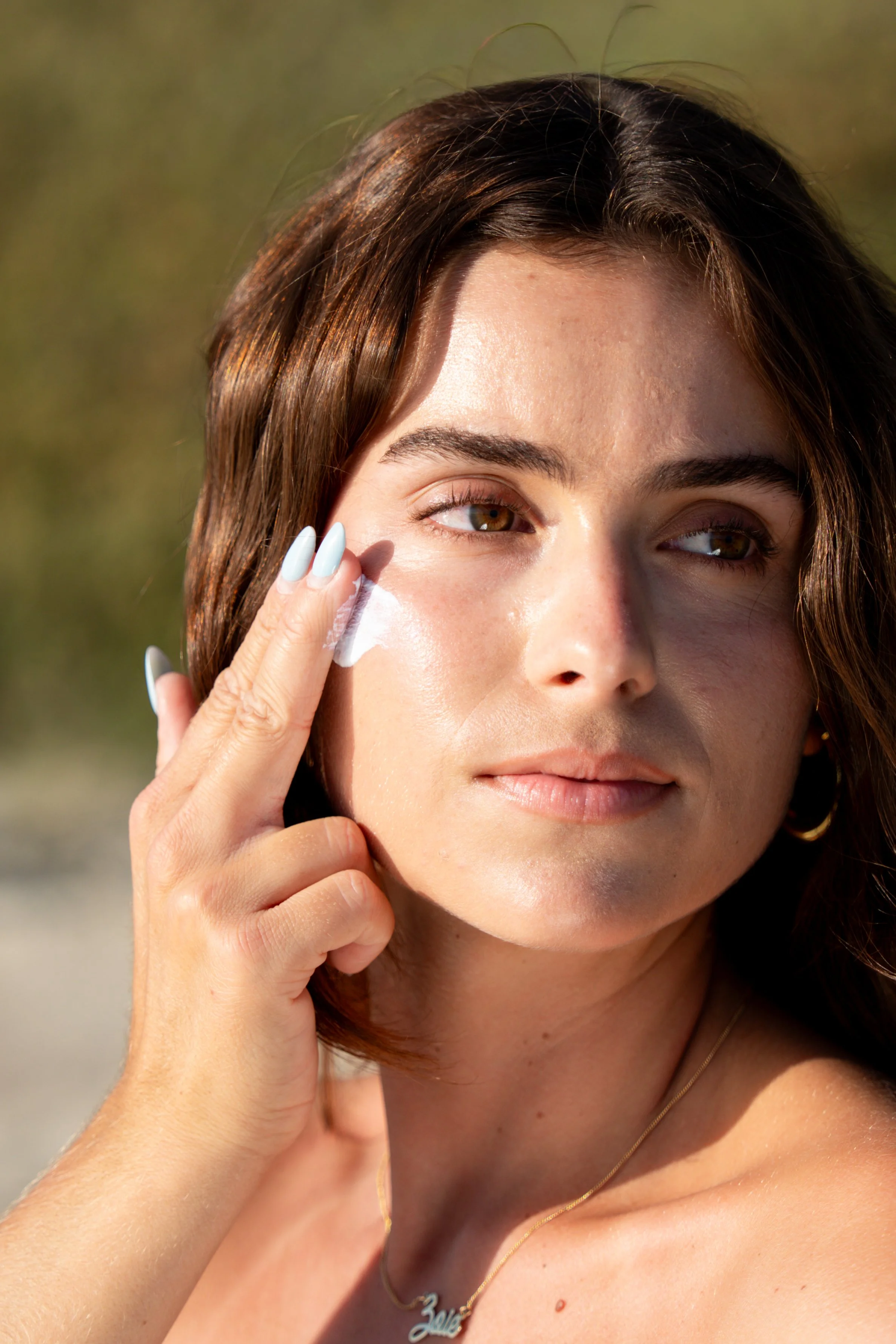 A young woman with brown hair and light skin applying sunscreen to her face outdoors on a sunny day.