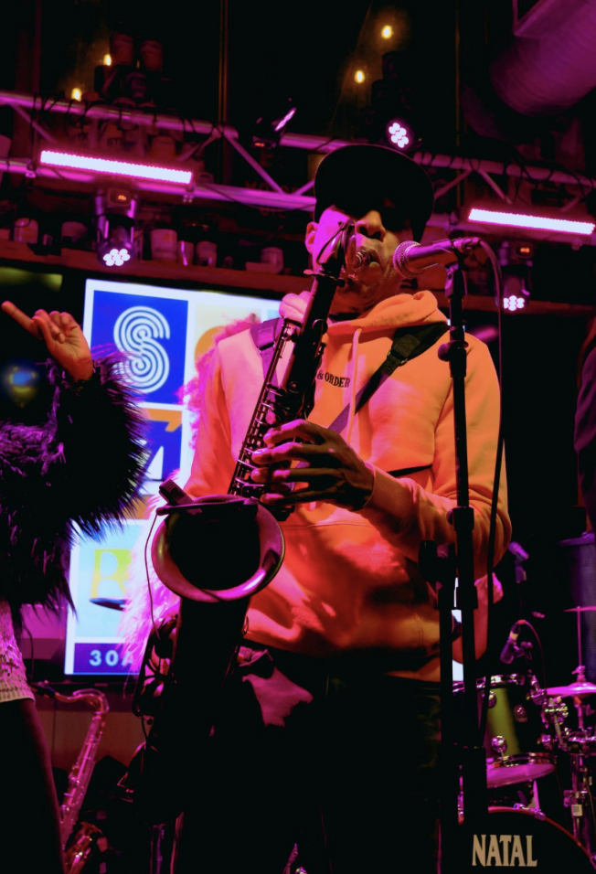 Musician playing saxophone on stage at night with colorful lights and a digital screen in the background.