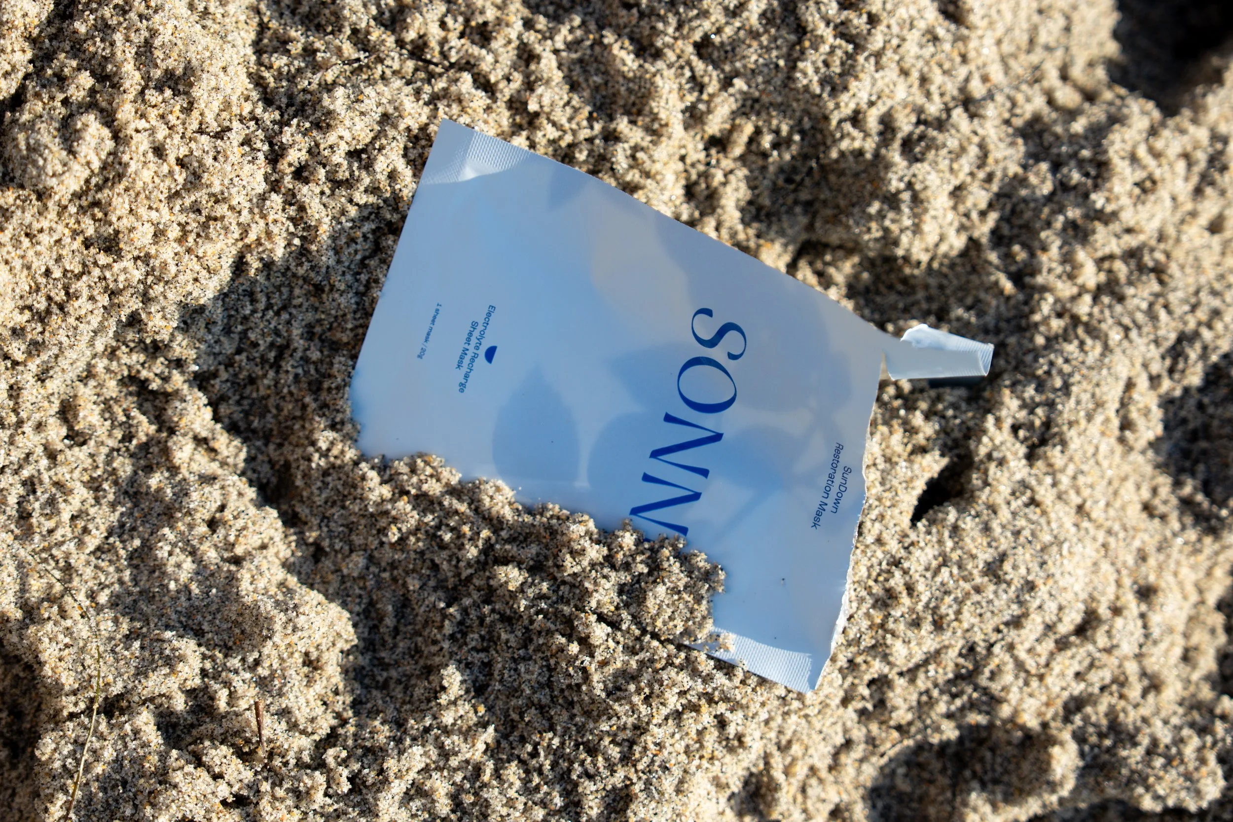 Sand with a torn open packet of a sheet mask lying on the beach, labeled 'Sand Vacation Sheet Mask'.