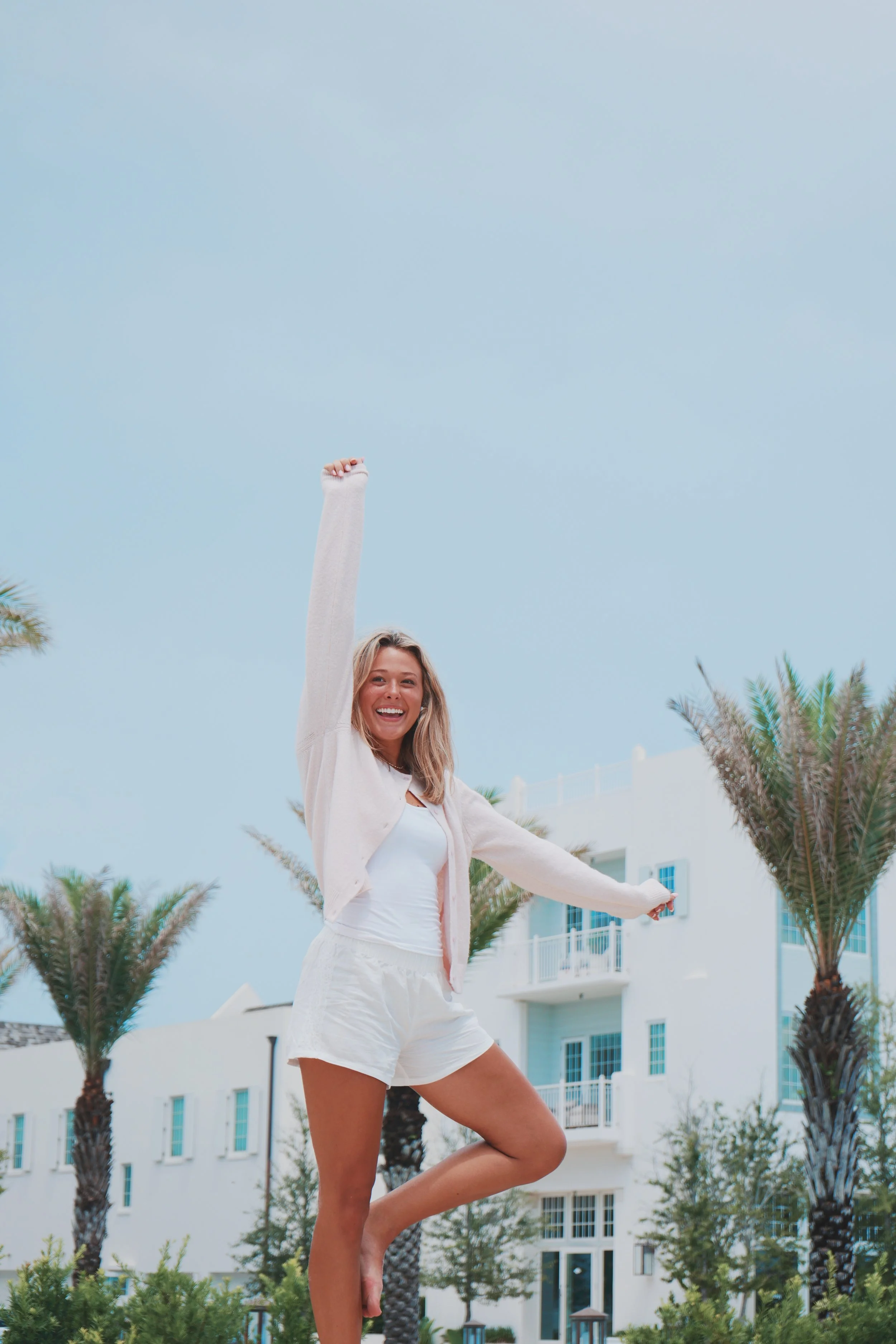 A young woman smiling and dancing outdoors near a white building with balconies, palm trees, and a bright blue sky.