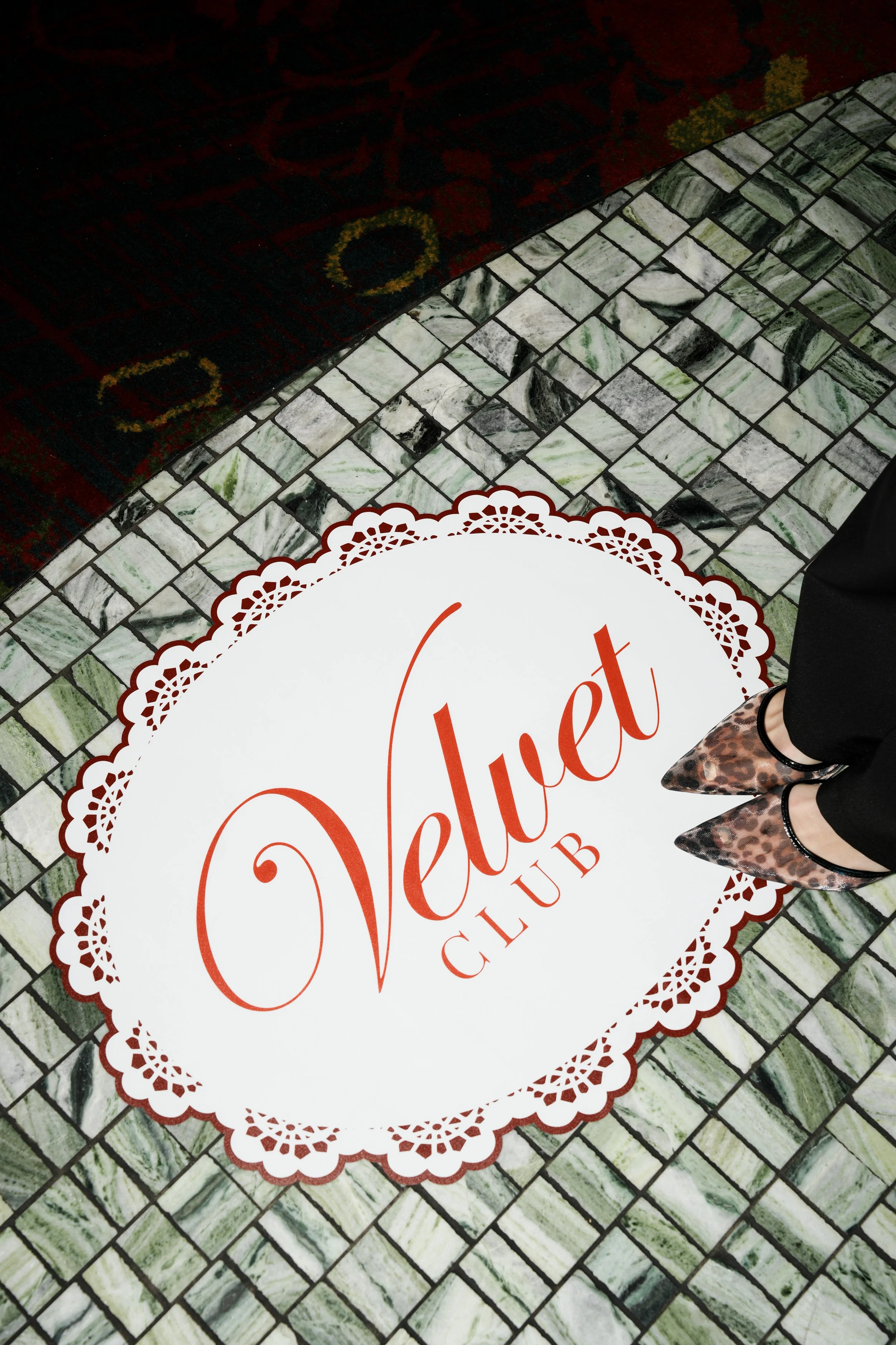 Part of a sign with the word "Violet" in cursive and a decorative border, placed on a floor with green and white marble tiles. Shoes of a person's feet are visible near the sign.