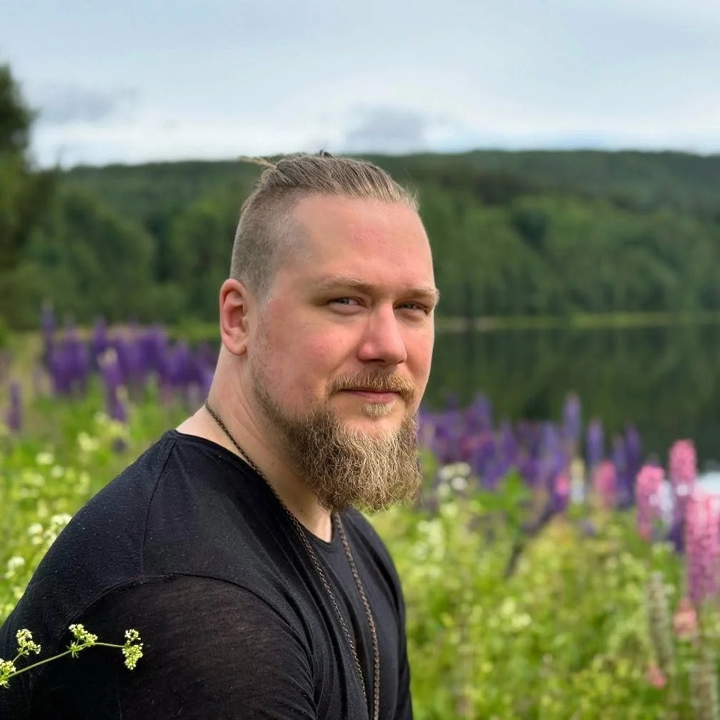 A man with a beard and styled hair wearing a black shirt, sitting outdoors near a lake with colorful flowers and green trees in the background.