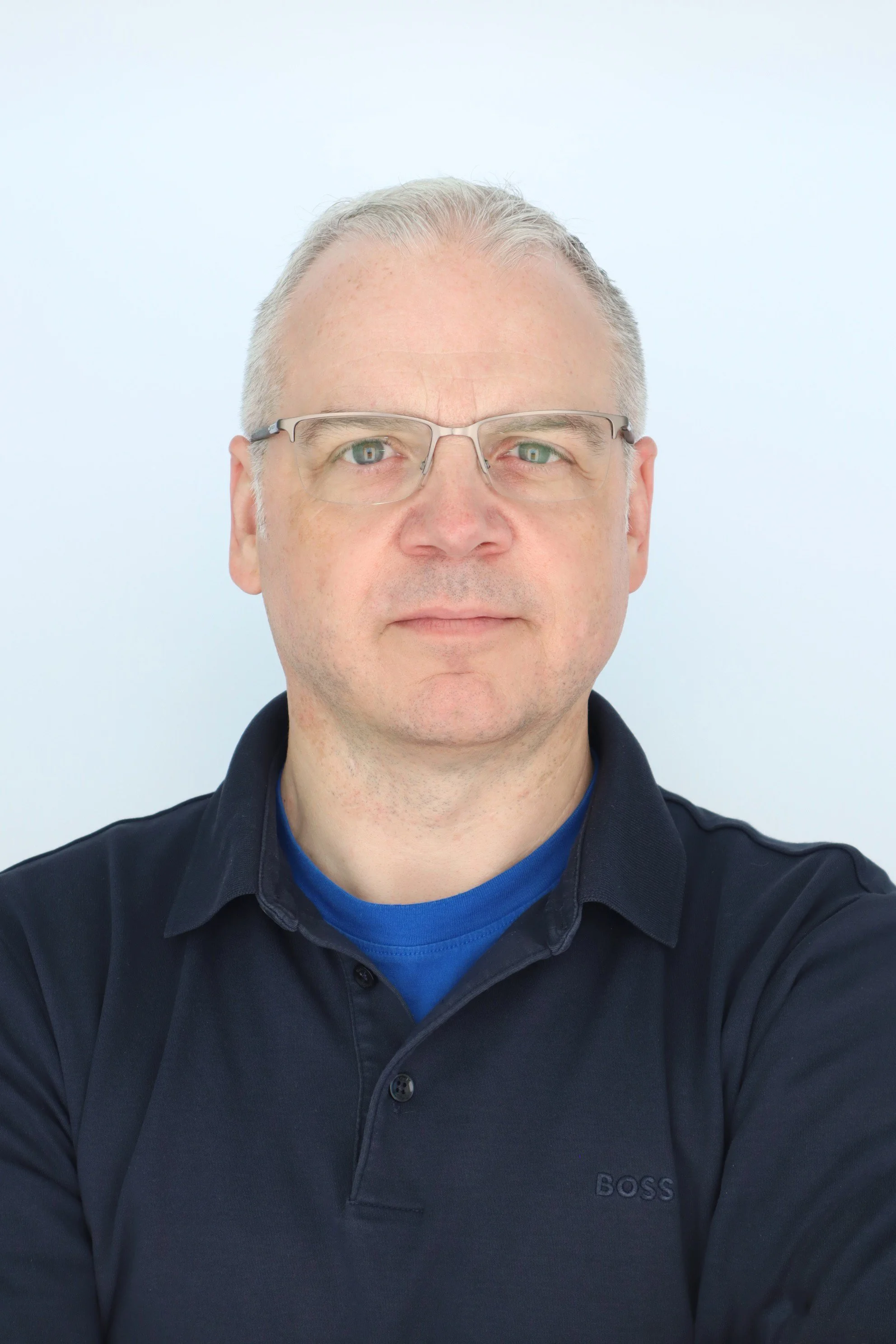 A middle-aged man with short gray hair, glasses, wearing a black long-sleeve shirt over a blue T-shirt, taking a selfie against a plain white background.