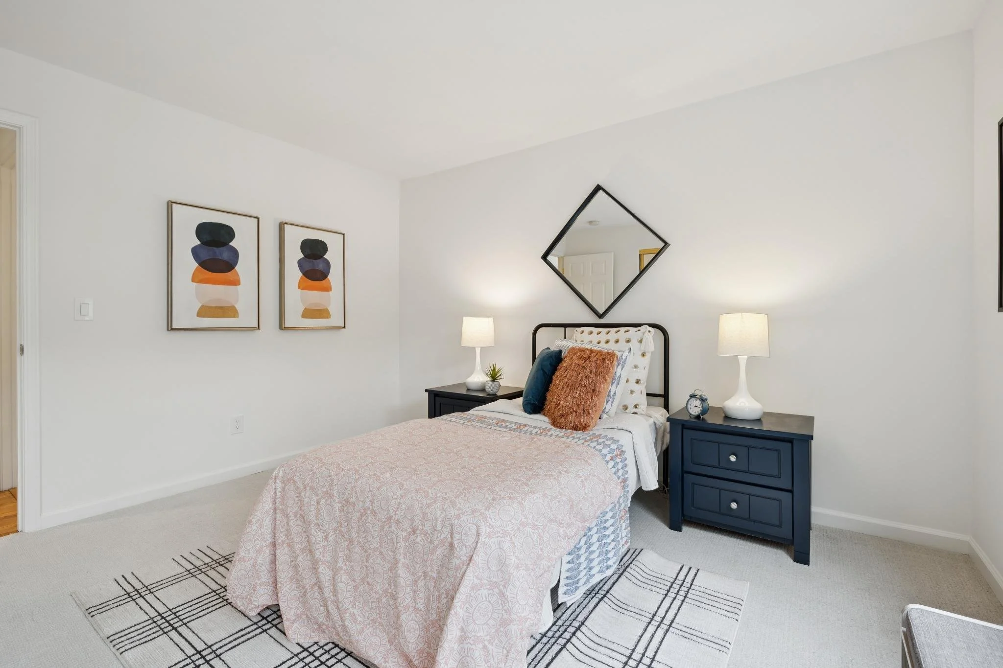 A bedroom with a bed covered in a pink patterned bedspread, decorative pillows, and a black metal headboard. Flanking the bed are two dark blue nightstands, each with a white table lamp. On one nightstand, there's a small plant and a clock. Above the