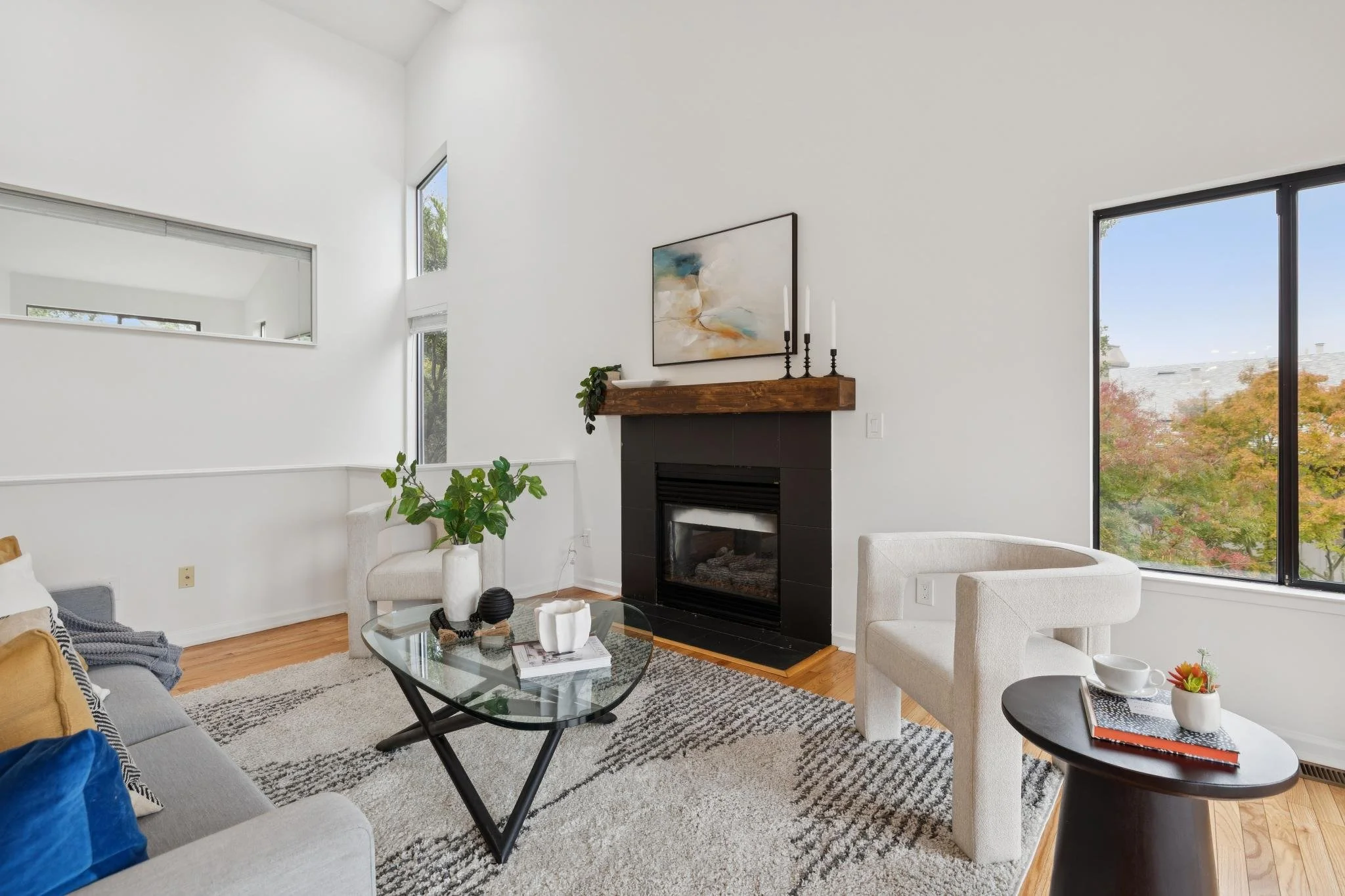 Modern living room with white walls, large windows showing trees, and a black fireplace with a wood mantel, decorated with candles and artwork.