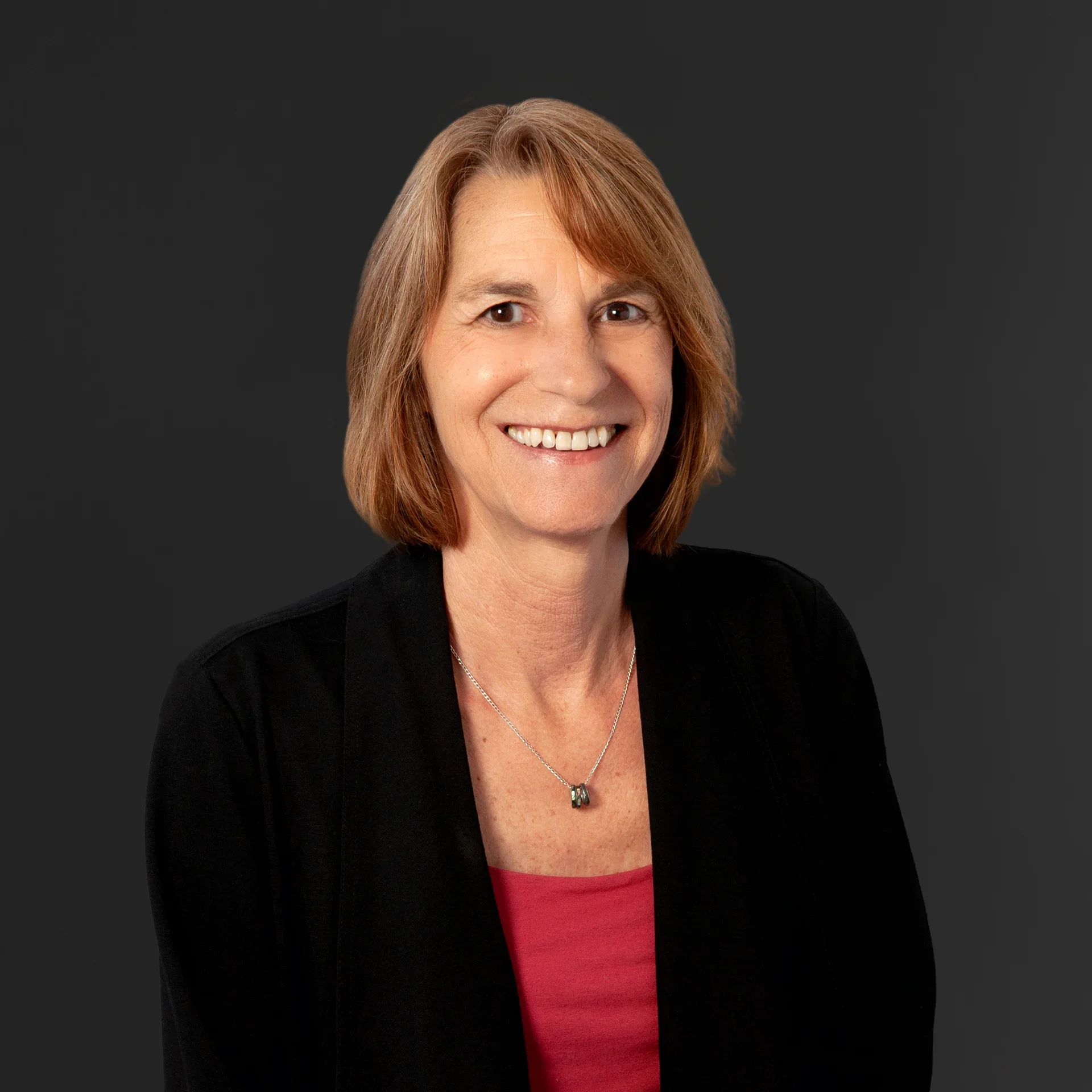 A woman with shoulder-length red hair, smiling, wearing a black jacket over a red top, and a silver necklace with a dark stone pendant, against a dark gray background.