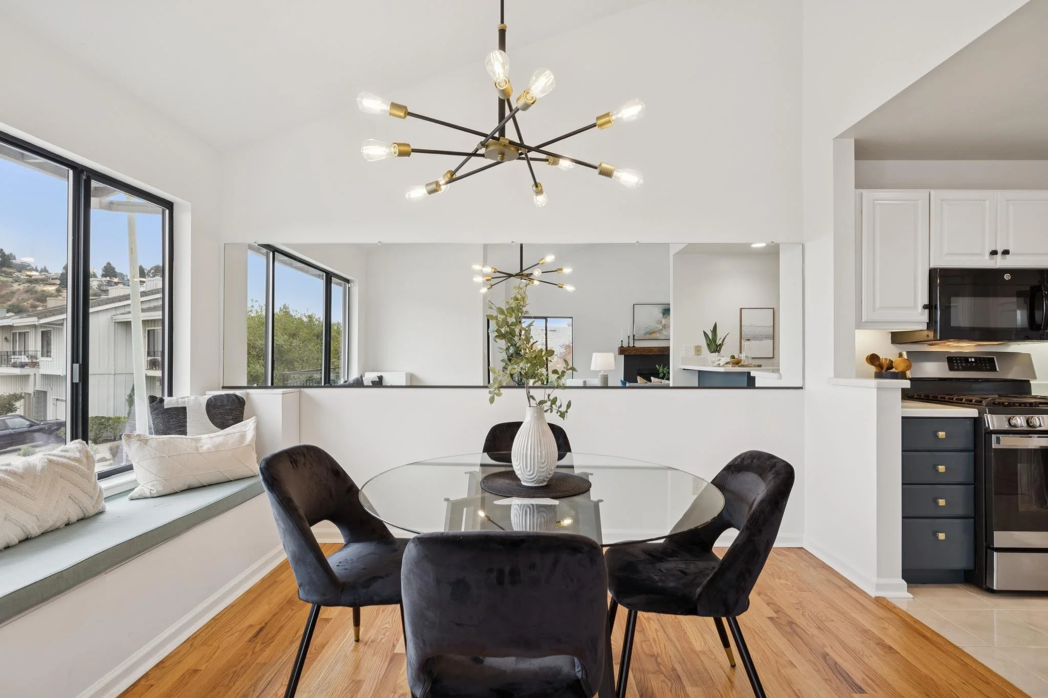 Modern dining area with black velvet chairs around a glass table, white walls, a large window, and a contemporary chandelier with multiple exposed bulbs.