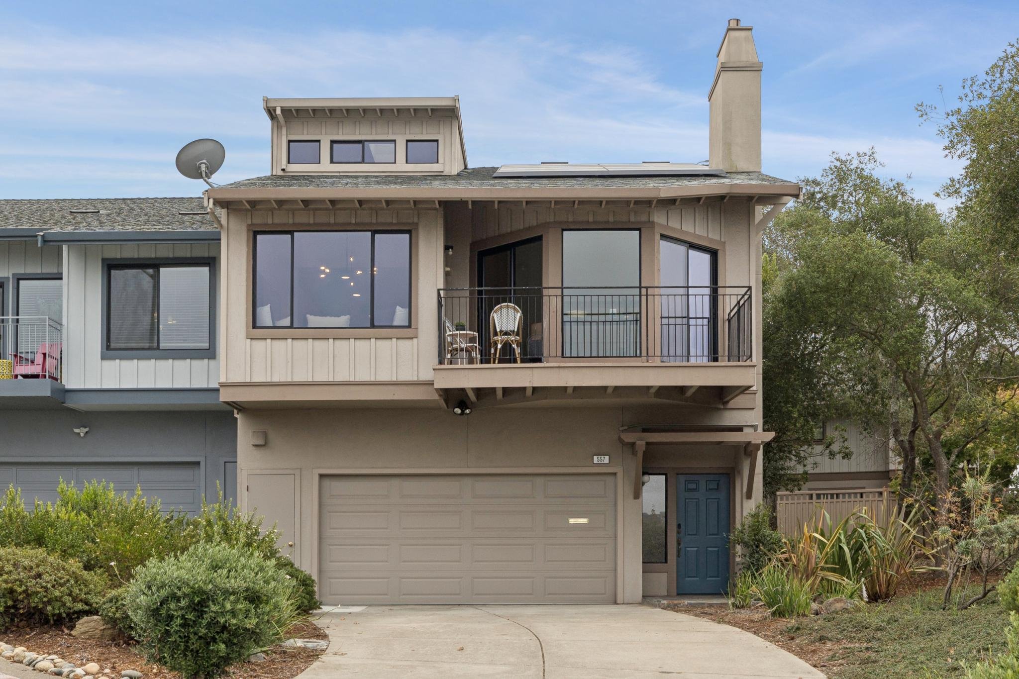 A modern multi-story house with a garage, balcony, and a chimney. The house has beige siding, large windows, and a blue front door. There are green bushes and trees in the front yard, and a driveway leading to the garage.