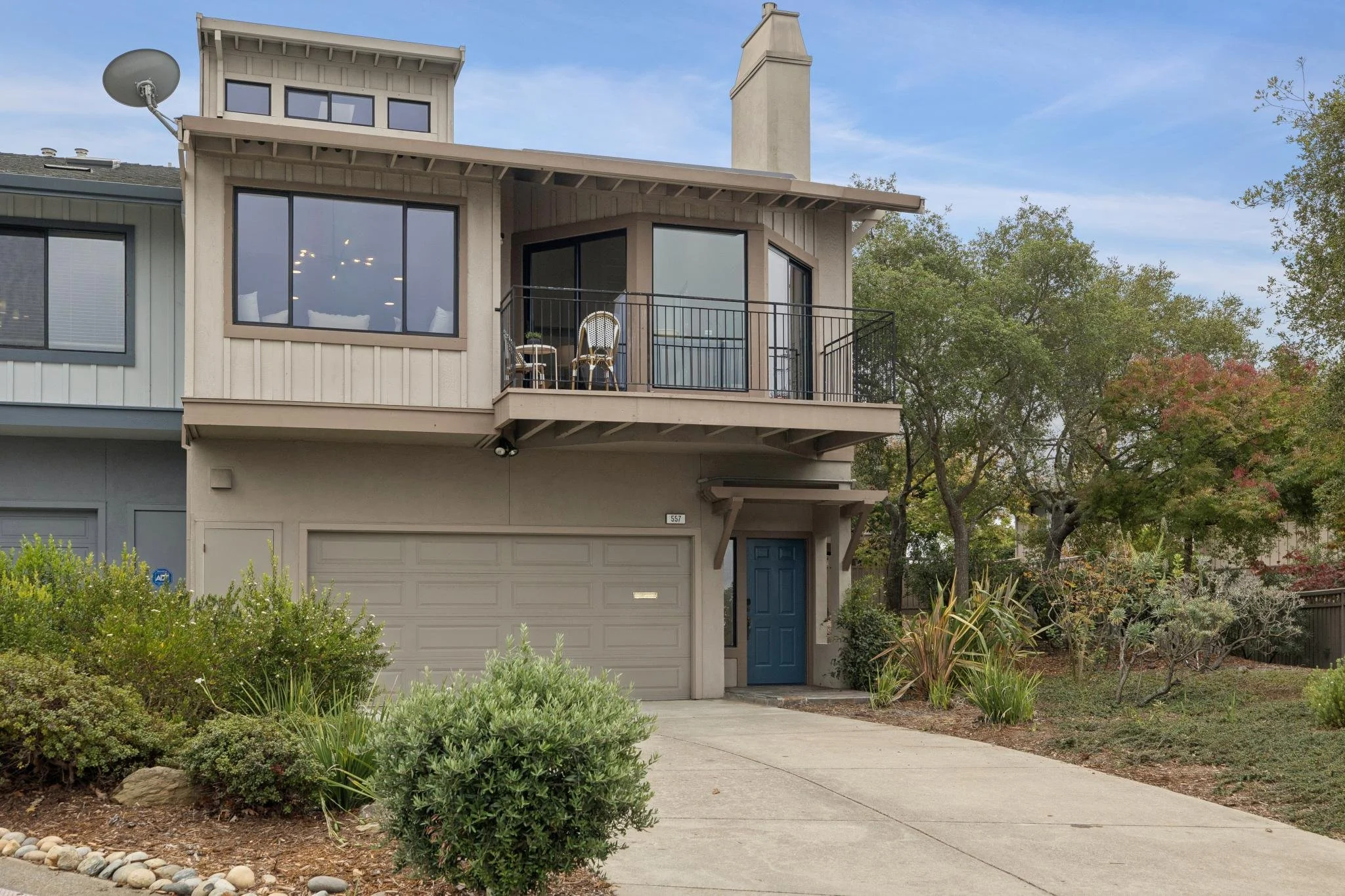 Front view of a modern two-story house with a garage, blue front door, balcony, large windows, and surrounding trees and bushes.