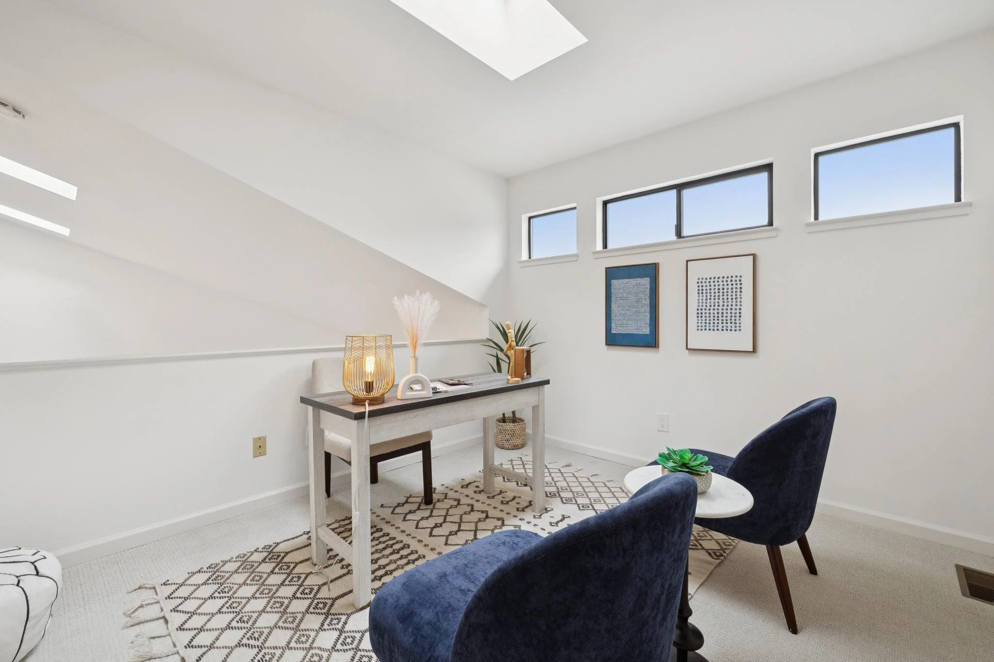 A cozy, minimalistic living space with white walls and high windows. Decor includes framed artwork, a table with a lamp, a plant, and a chair.