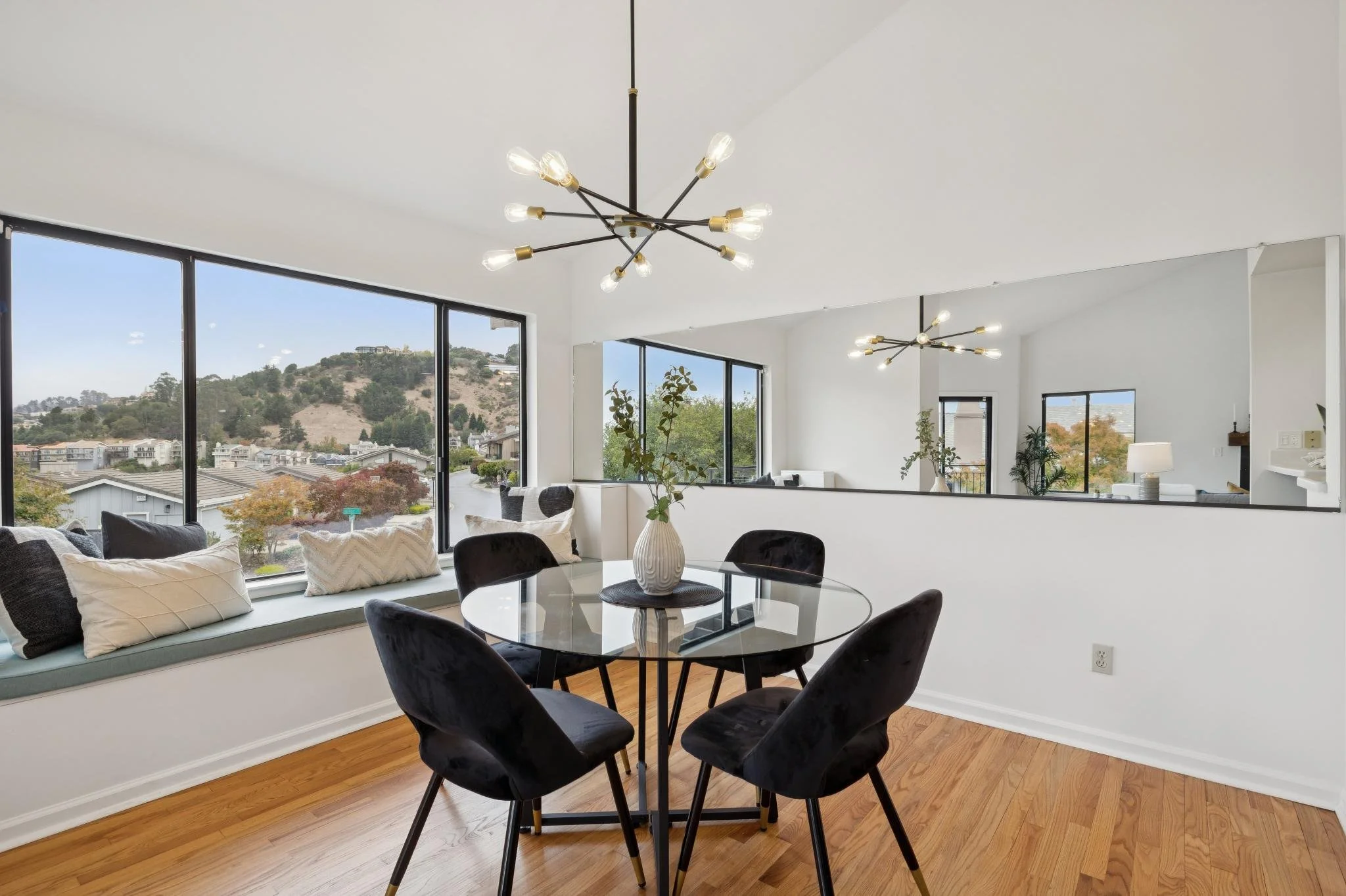 Modern dining room with black chairs around a round glass table, white walls, large windows showing hillside views, and contemporary light fixtures.