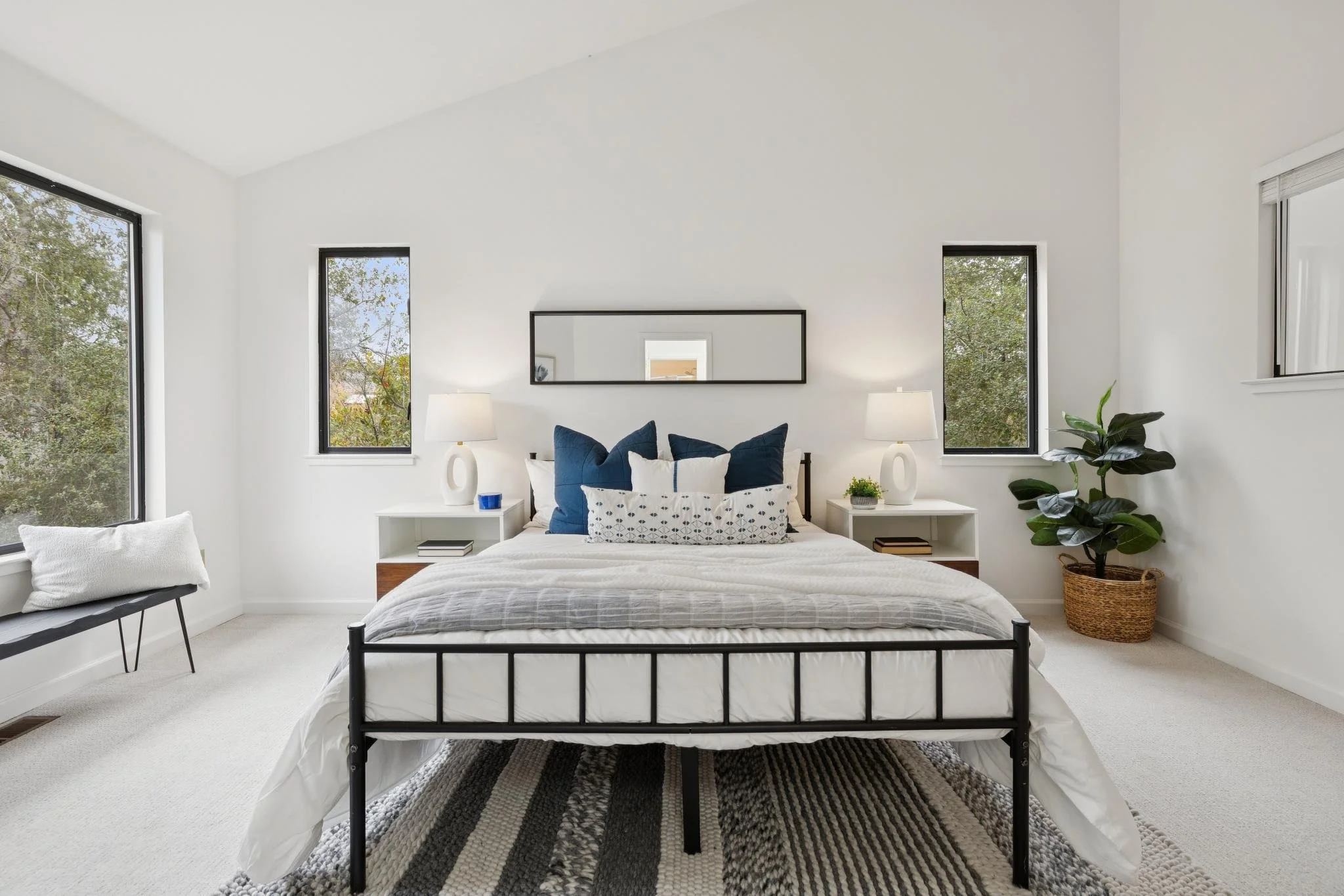 A modern bedroom with large windows, white walls, a black metal bed frame, white bedding, and blue accent pillows. There are two white nightstands with lamps, a mirror above the bed, a potted plant on the right, and a black and white striped rug on t