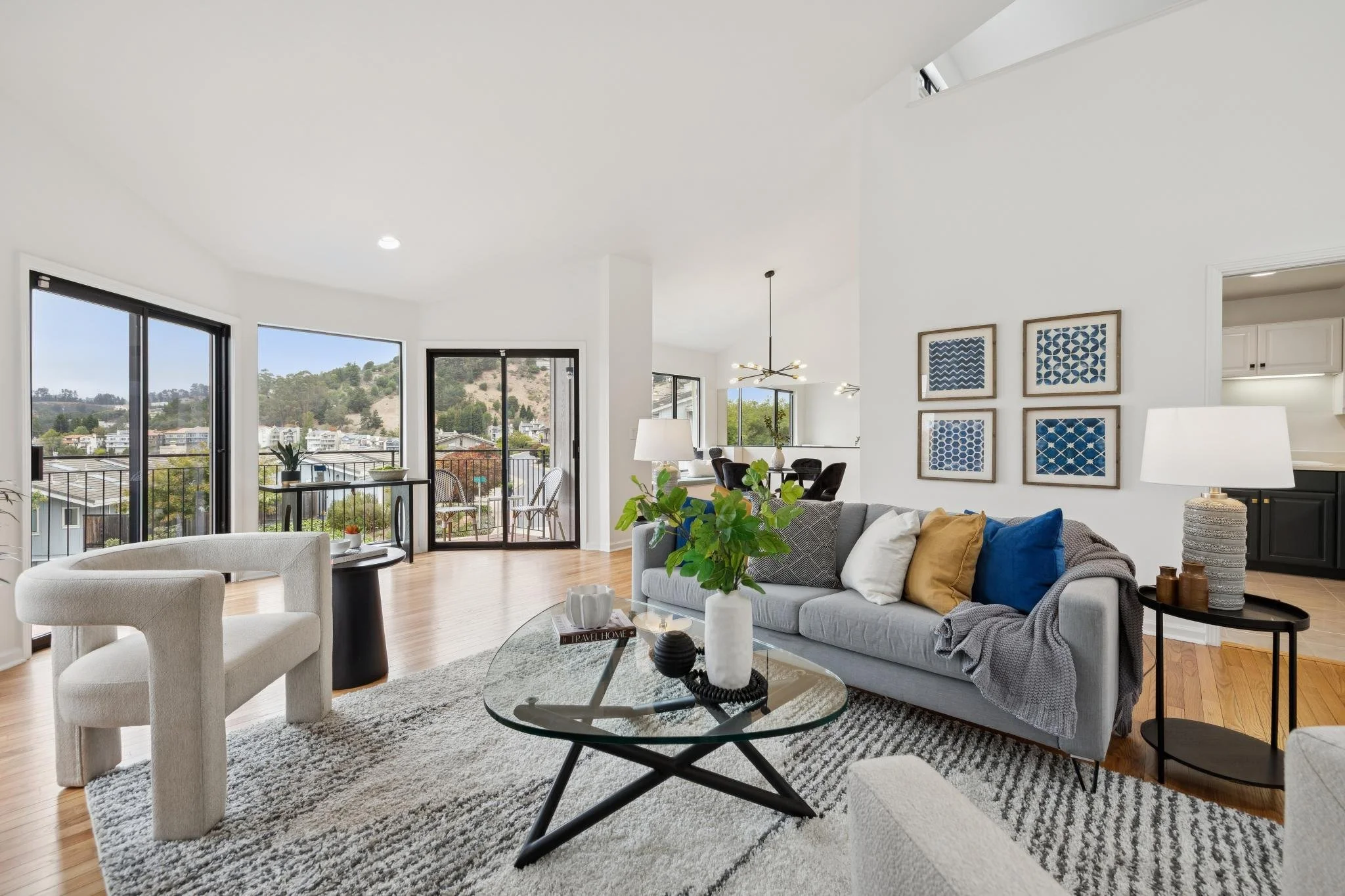 Living room with large windows showing hillside scenery, modern furniture, a gray sofa with pillows, a glass coffee table, and wall art with blue patterns.