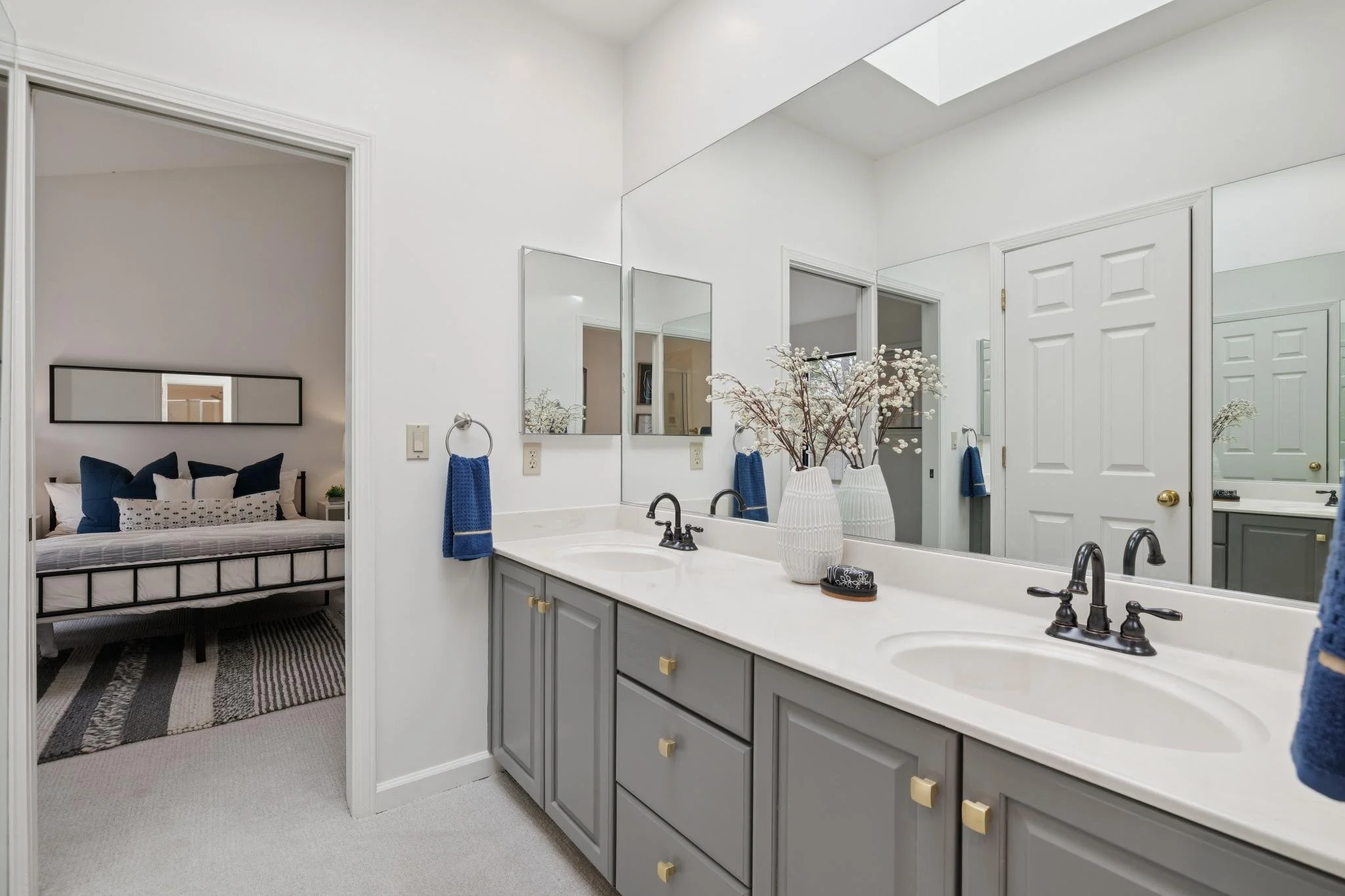 Bathroom with gray cabinets, a white countertop, and a large mirror. Decor includes white vases with white flowers, blue towels, and a soap dish. Connected to a bedroom with a bed, navy pillows, mirror, and a striped rug.